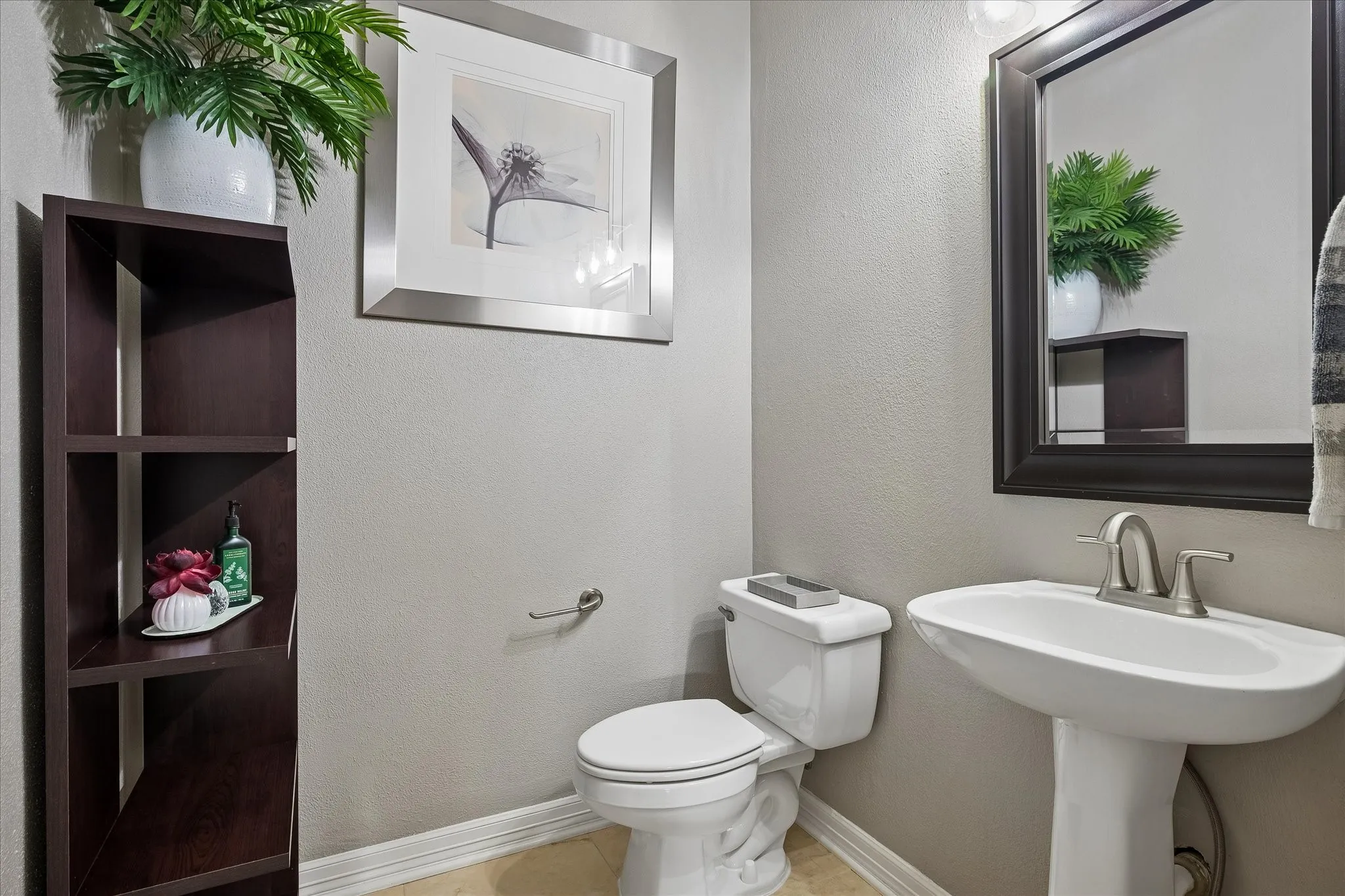 Pedestal sink with a brushed nickel faucet, large framed mirror, wood-finish shelving unit, and tile flooring