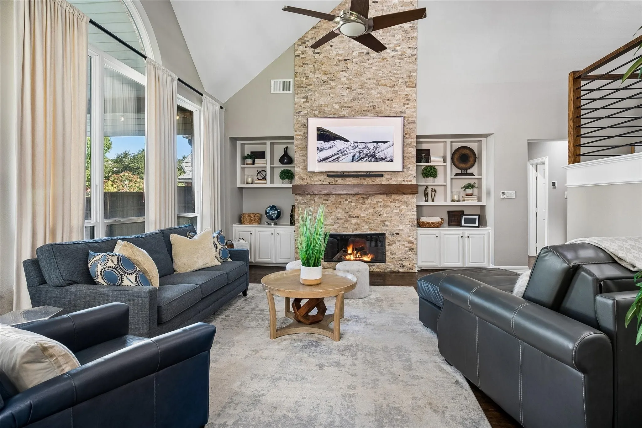 Vaulted ceiling with a ceiling fan, stone-clad fireplace with a wood mantle, built-in shelving and cabinetry, and large arched windows
