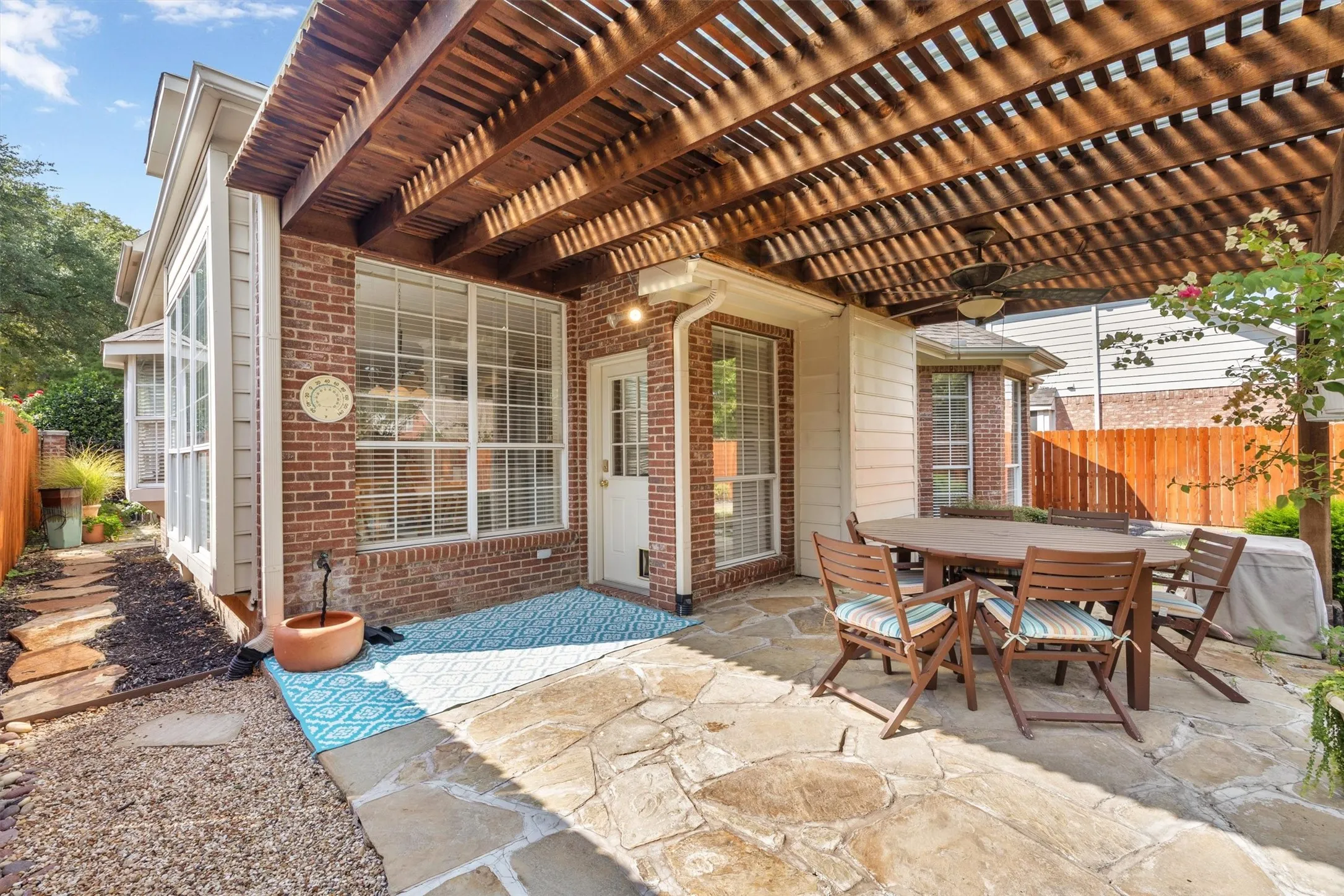 Expansive stone patio with an overhead wooden pergola and ceiling fan, complemented by brick and siding exterior walls, and a private wooden fence