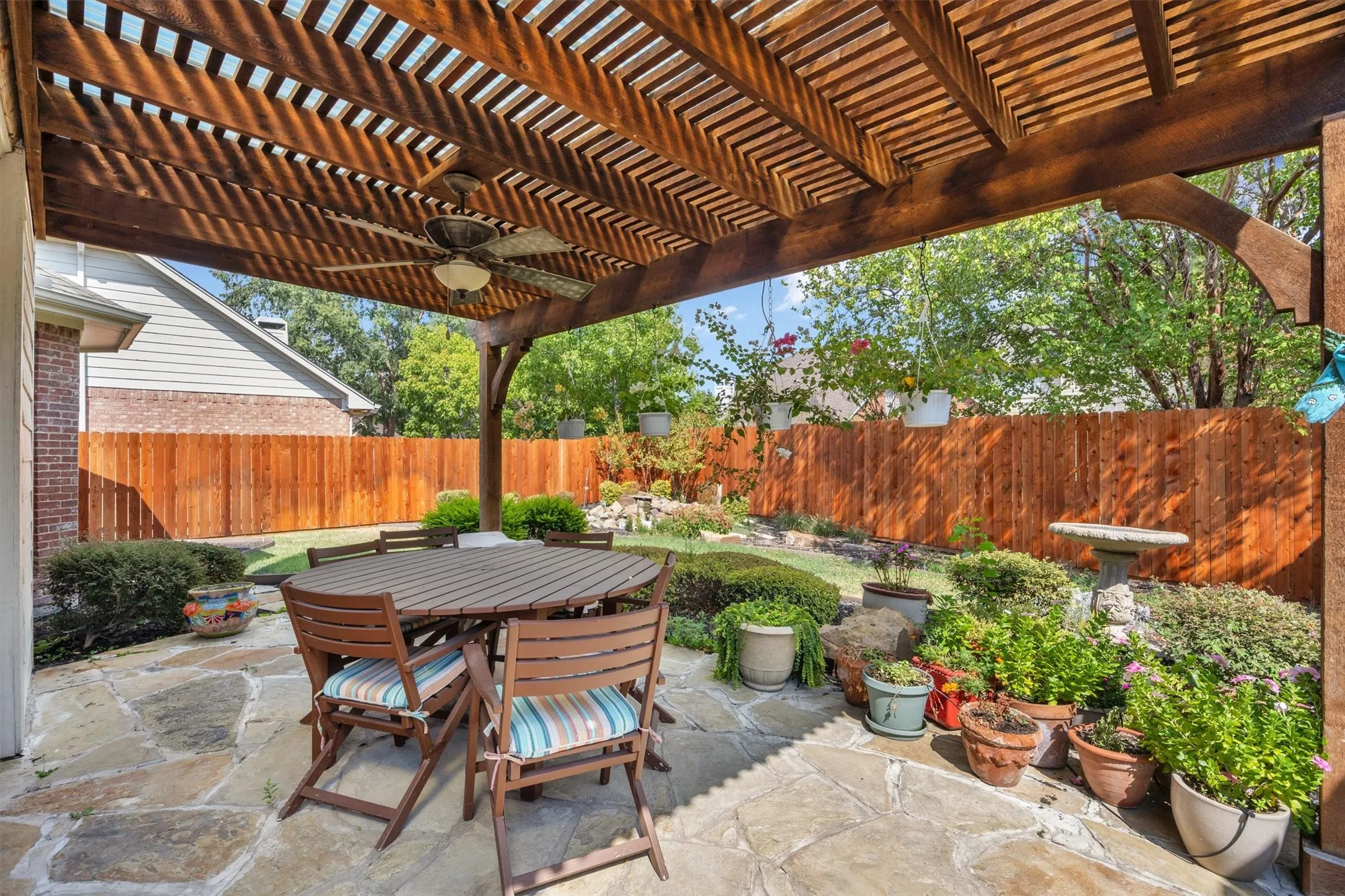 Expansive flagstone patio featuring a wood pergola with an integrated ceiling fan, surrounded by a wood privacy fence and landscaped garden beds