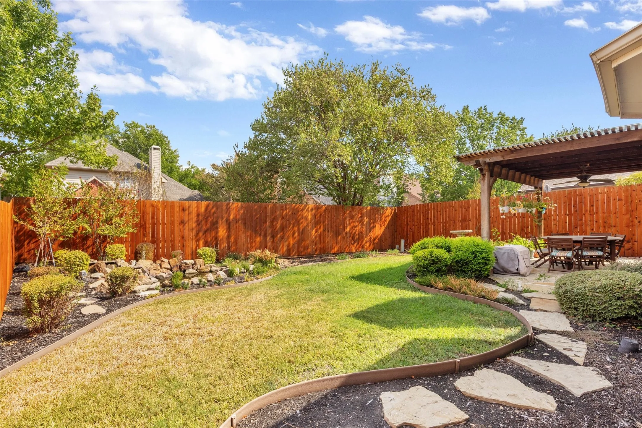 Expansive backyard featuring a stained wood privacy fence, a natural stone-edged lawn, a rock garden, and a wooden pergola covering a patio area with irregular stone pavers