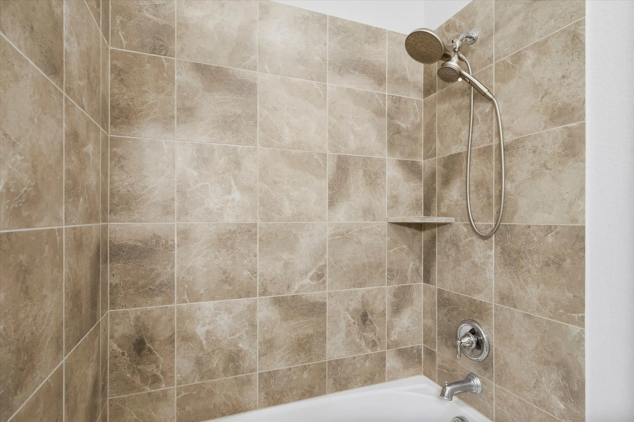 Bathtub and shower combination featuring a brushed nickel showerhead and faucet, tiled walls, and a built-in corner shelf