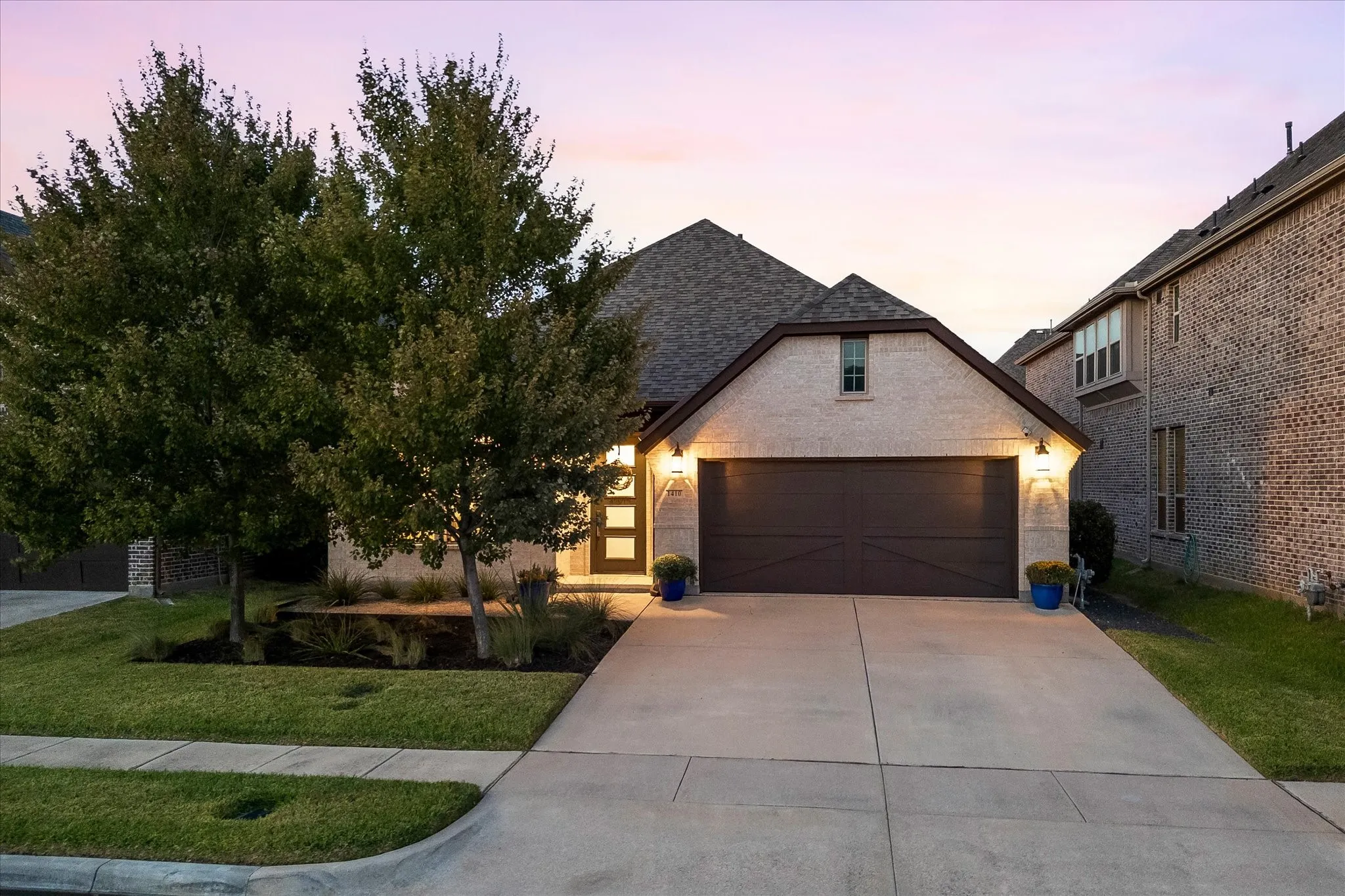 Light-colored brick exterior with a contrasting dark brown garage door, front landscaping with mature trees, a concrete driveway, and a gabled roofline