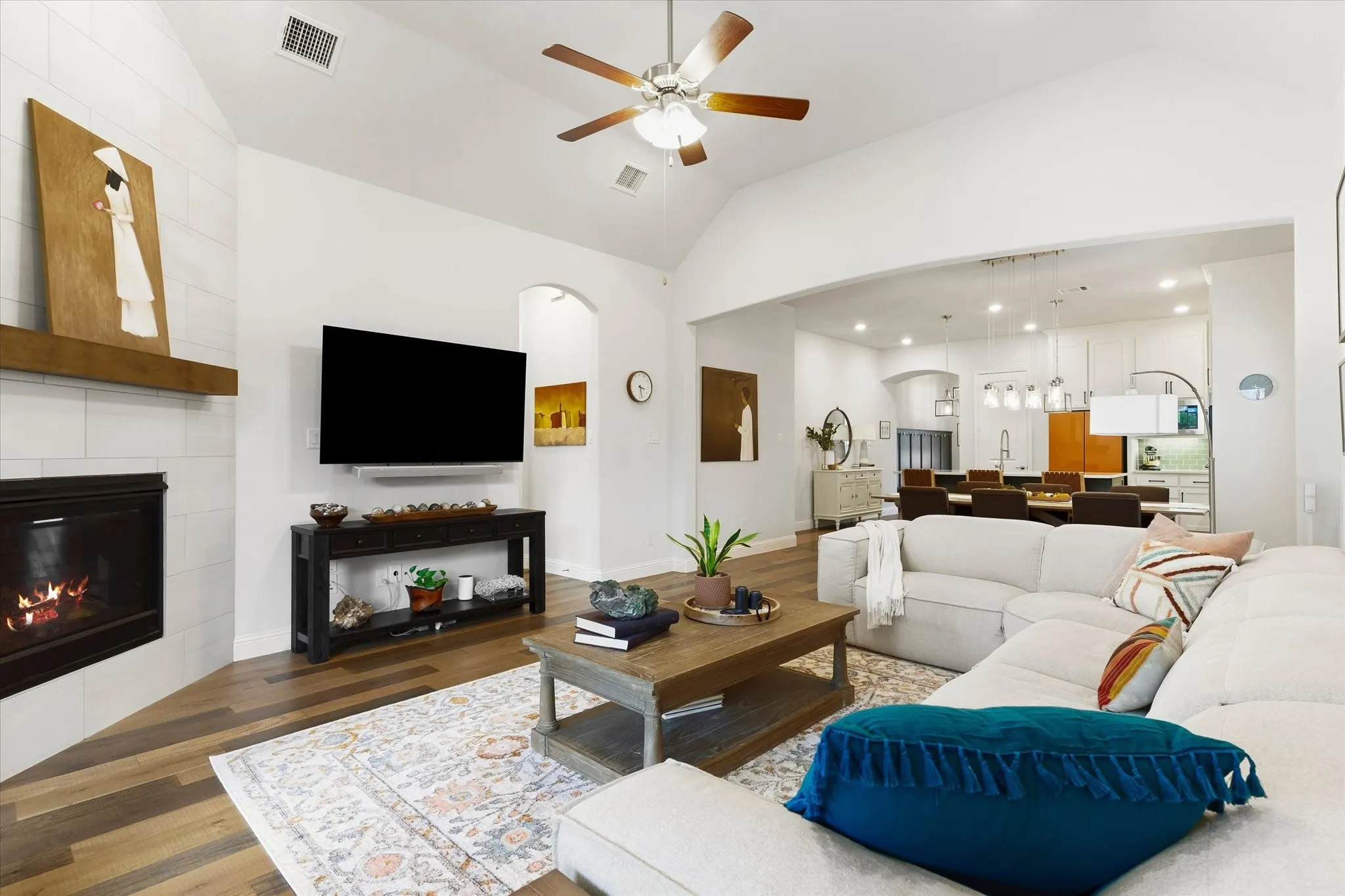Vaulted ceiling with a ceiling fan, fireplace with a tile surround and wood mantel, wood-finish flooring, and an arched entry to the adjacent room