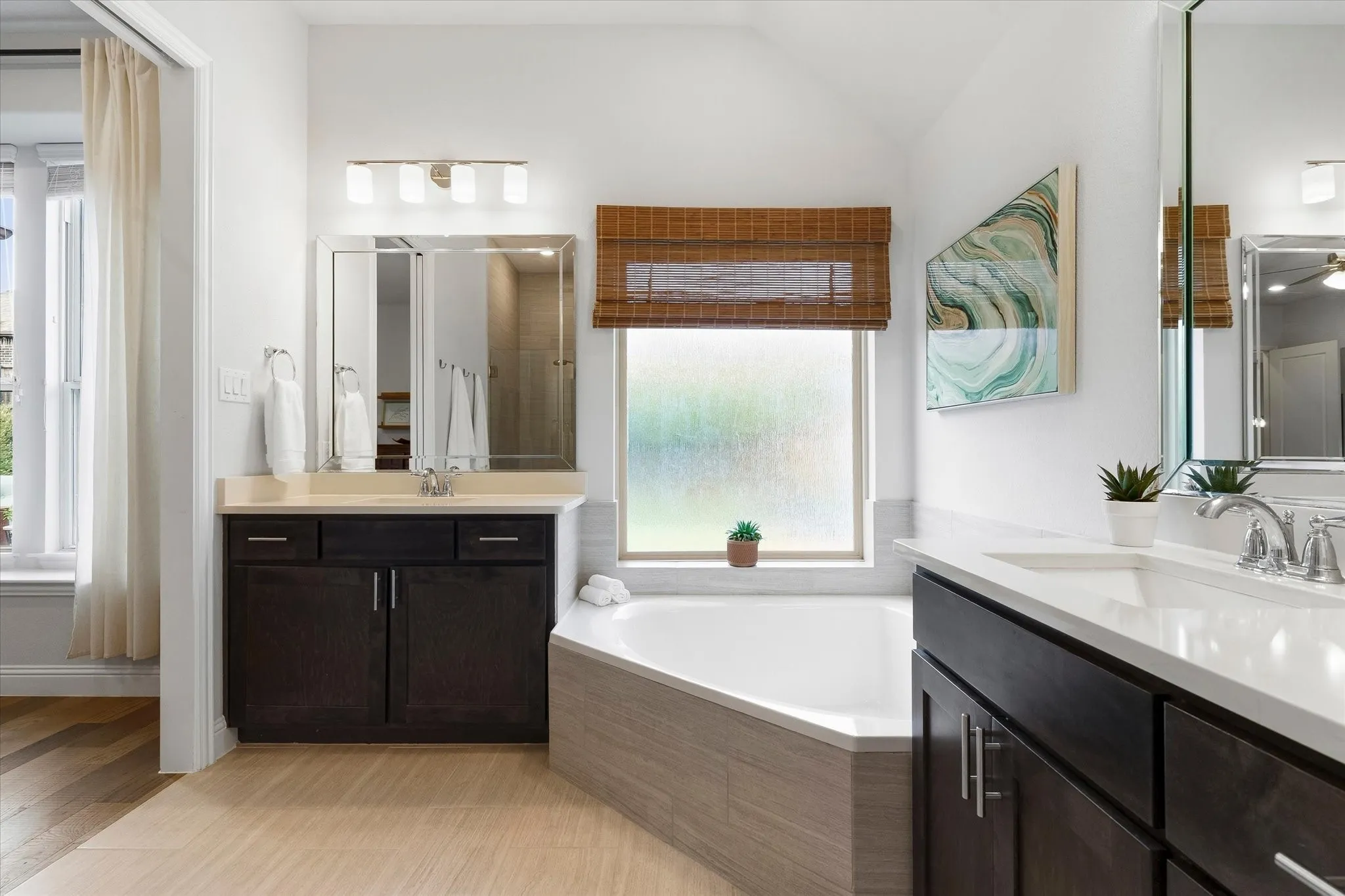 Dual vanities with undermount sinks, corner soaking tub with tiled surround, wood-finish flooring, and a window with a woven shade