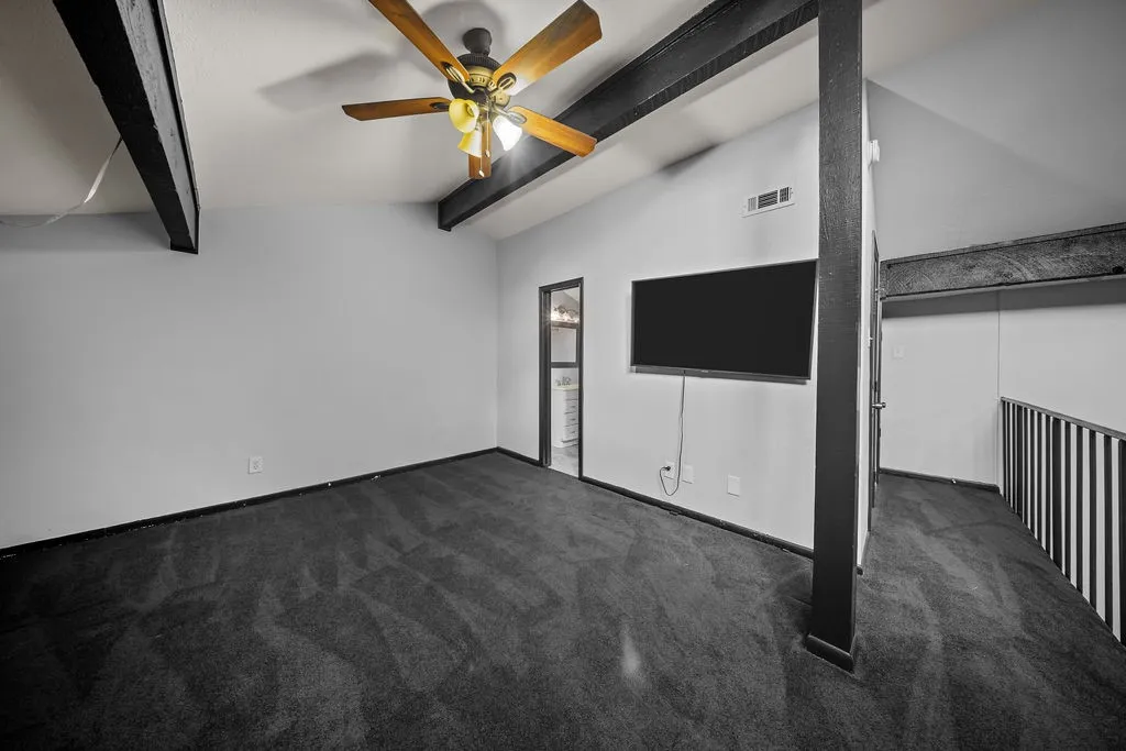 Vaulted ceiling with exposed beams, wood-finish ceiling fan, wall-mounted television, and dark carpeting