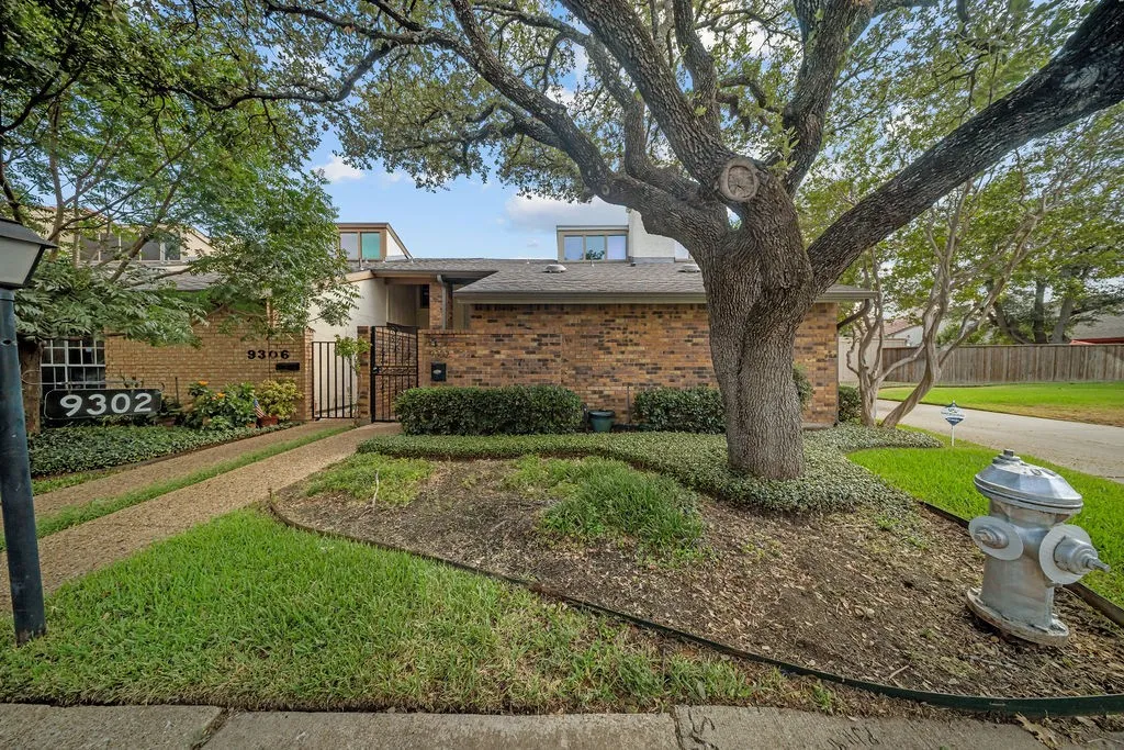 Brick facade residence with a mature front yard tree, a gravel pathway, and an ornamental security gate