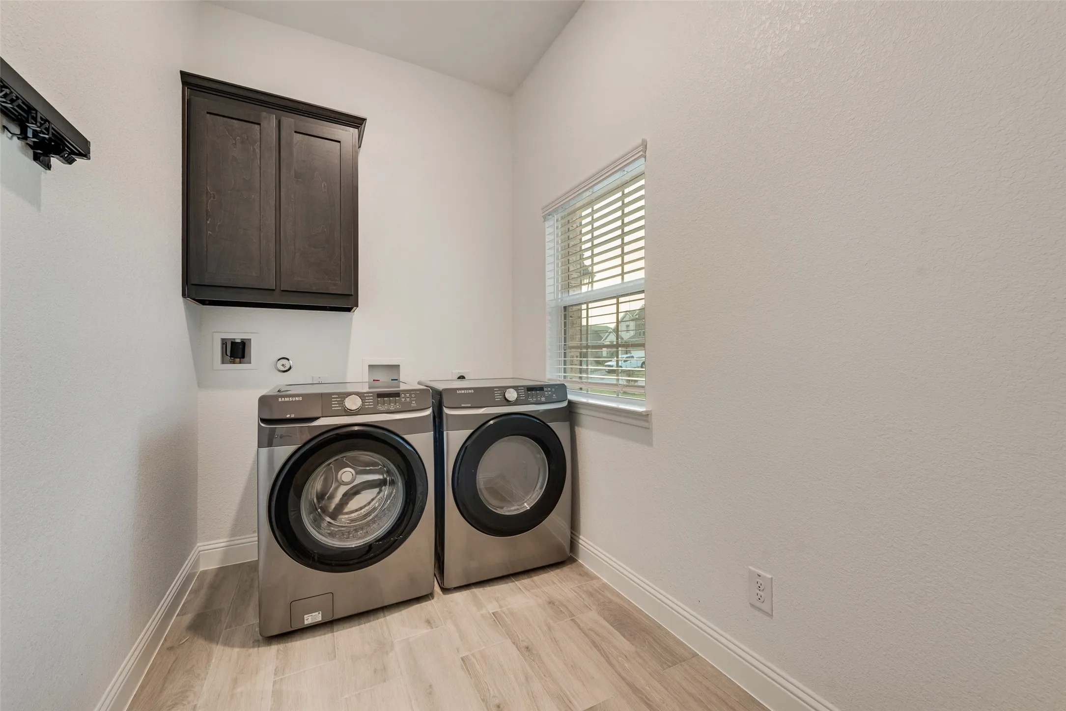 Laundry room featuring wood-finish flooring, a built-in dark wood-tone cabinet, and a window with blinds
