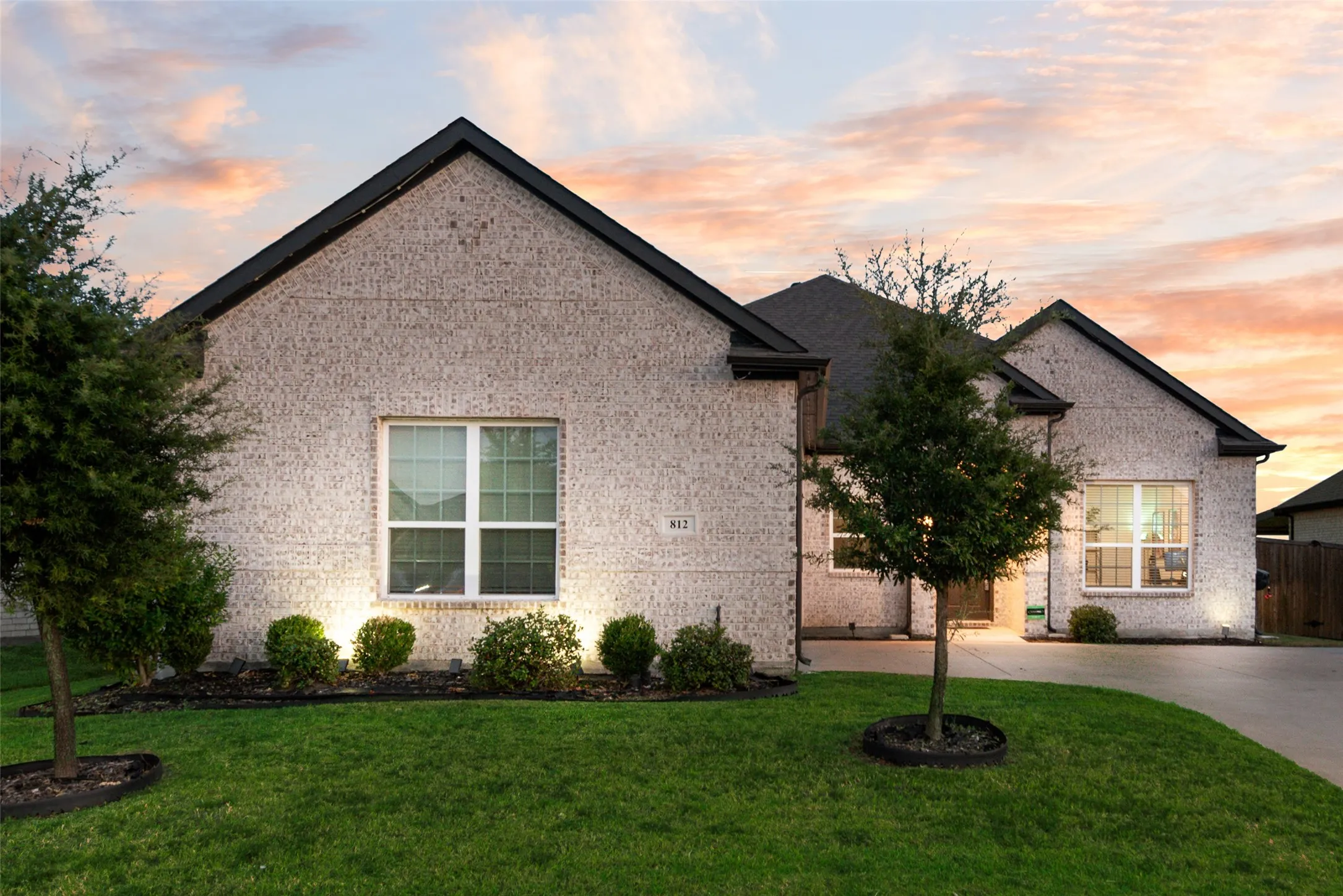White brick exterior, black shingle roof, integrated landscaping with shrubs and trees, manicured lawn, and paved driveway