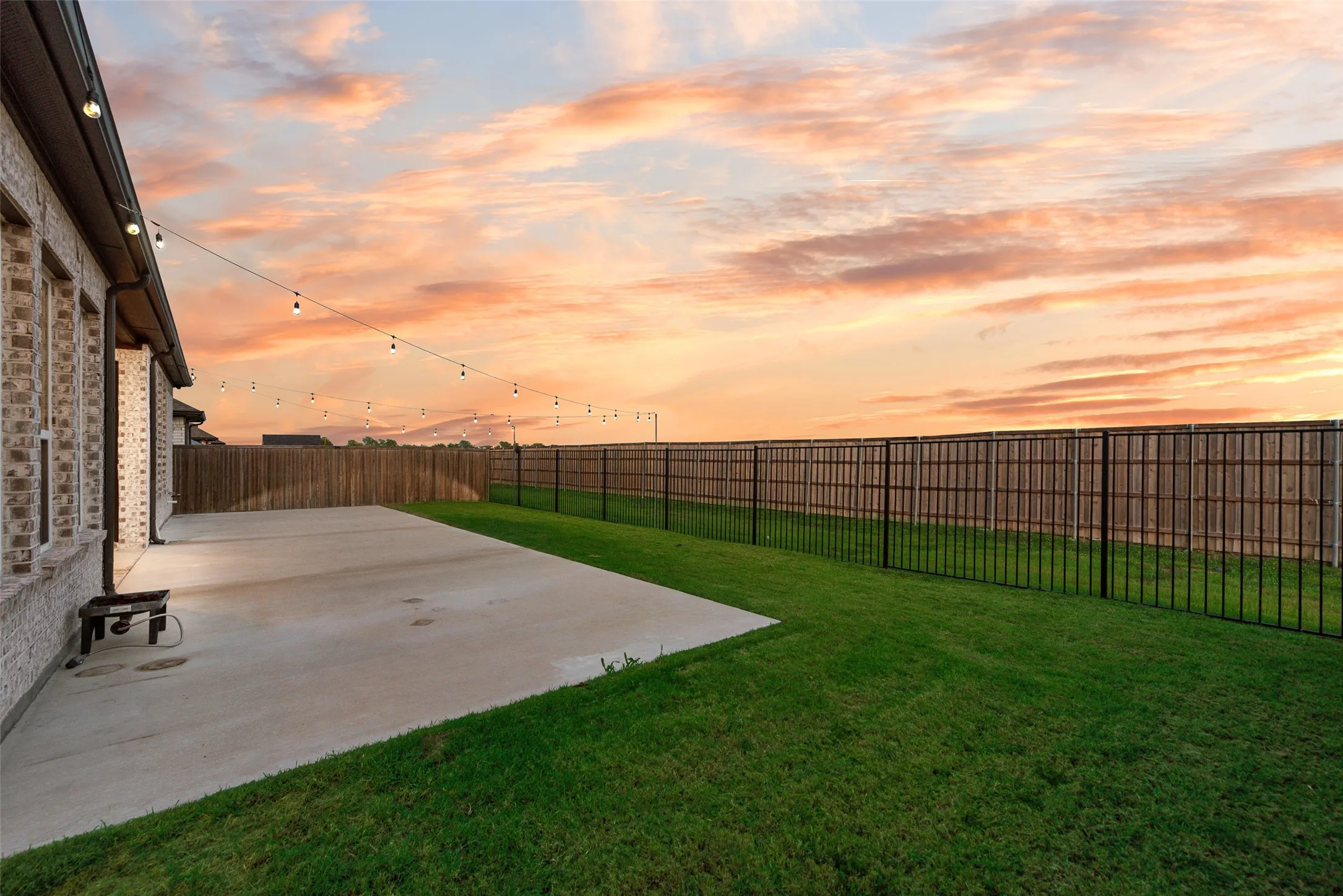 Expansive concrete patio with string lighting, bordered by a green lawn and a combination wood and metal fence