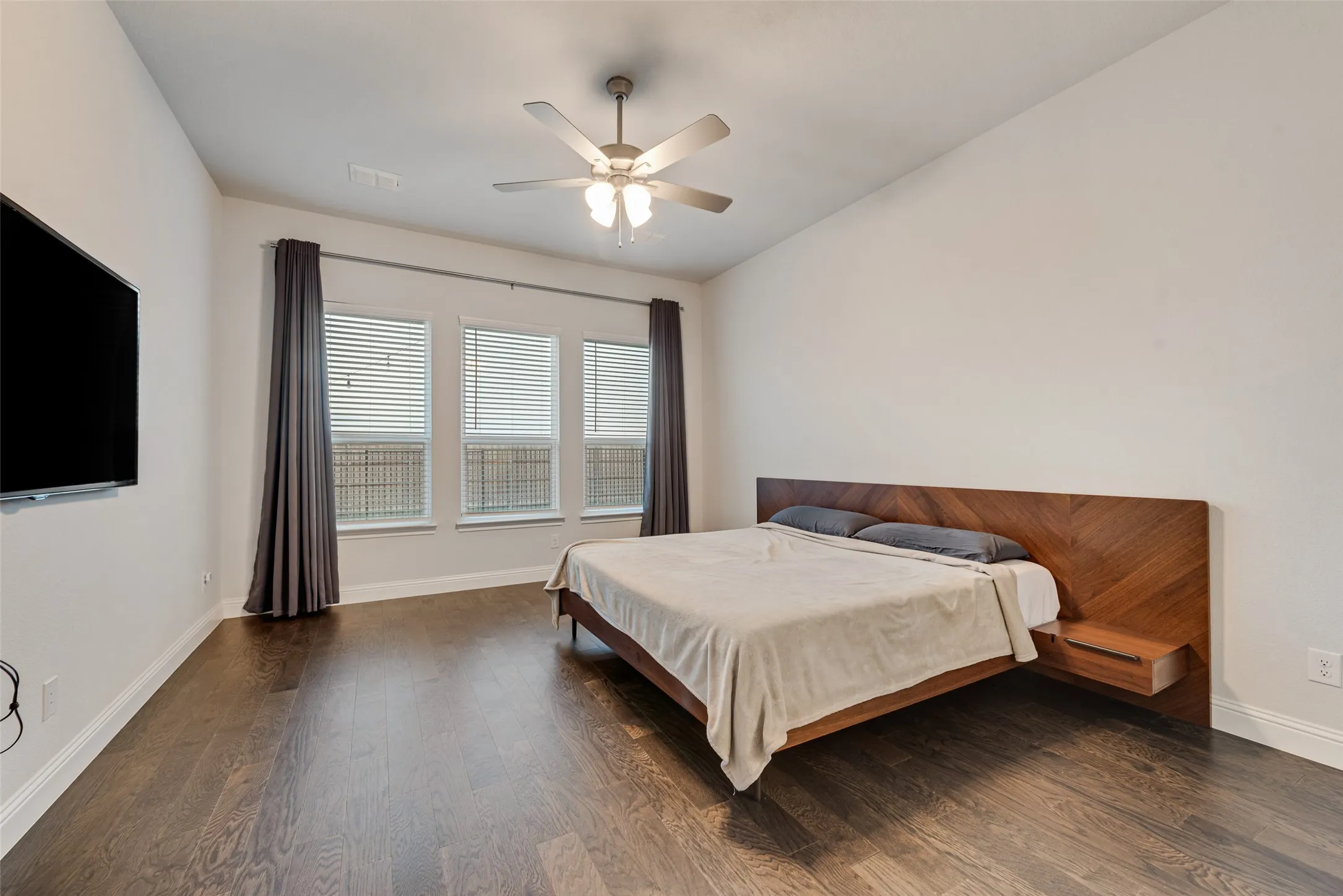 Dark wood-finish flooring, three large windows, and a ceiling fan with integrated lighting
