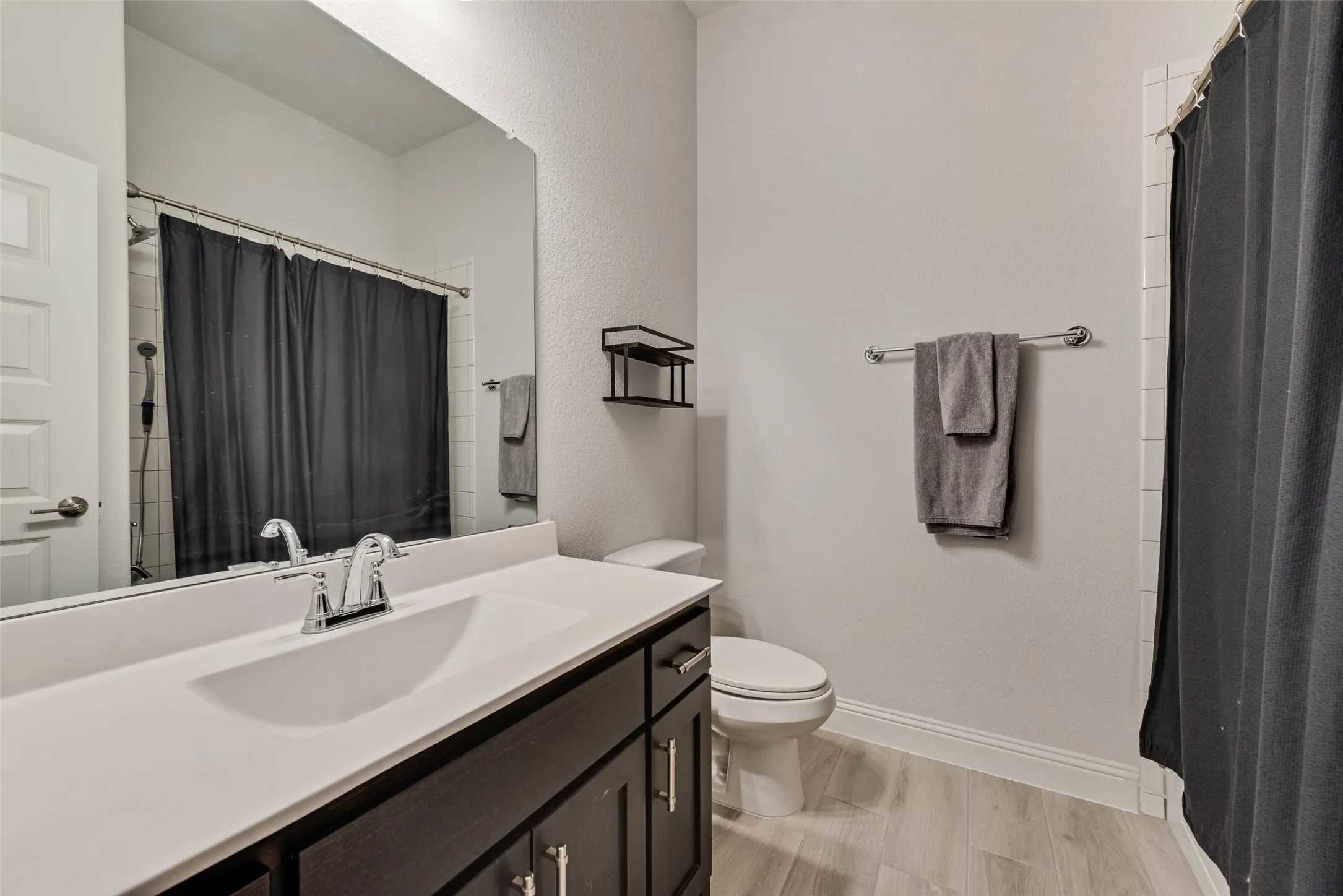 Bathroom featuring a dark wood-finish vanity with a white countertop and integrated sink, a large wall-mounted mirror, light-toned wood-finish flooring, and a shower with white subway tile