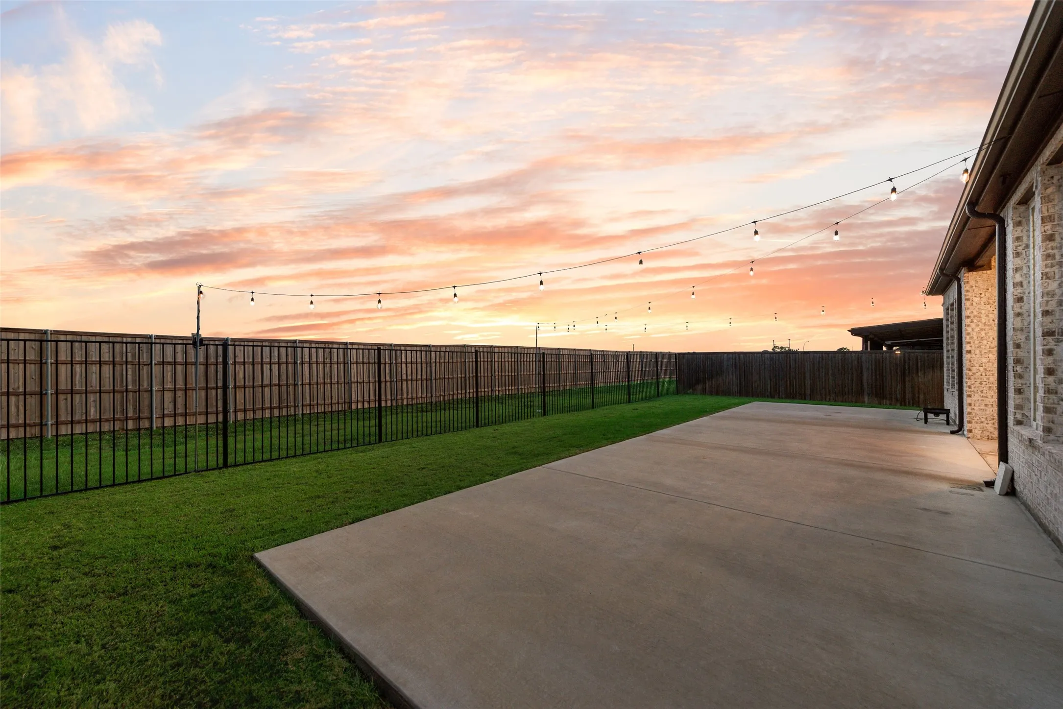 Expansive concrete patio with a manicured lawn, full privacy fencing, and string lighting