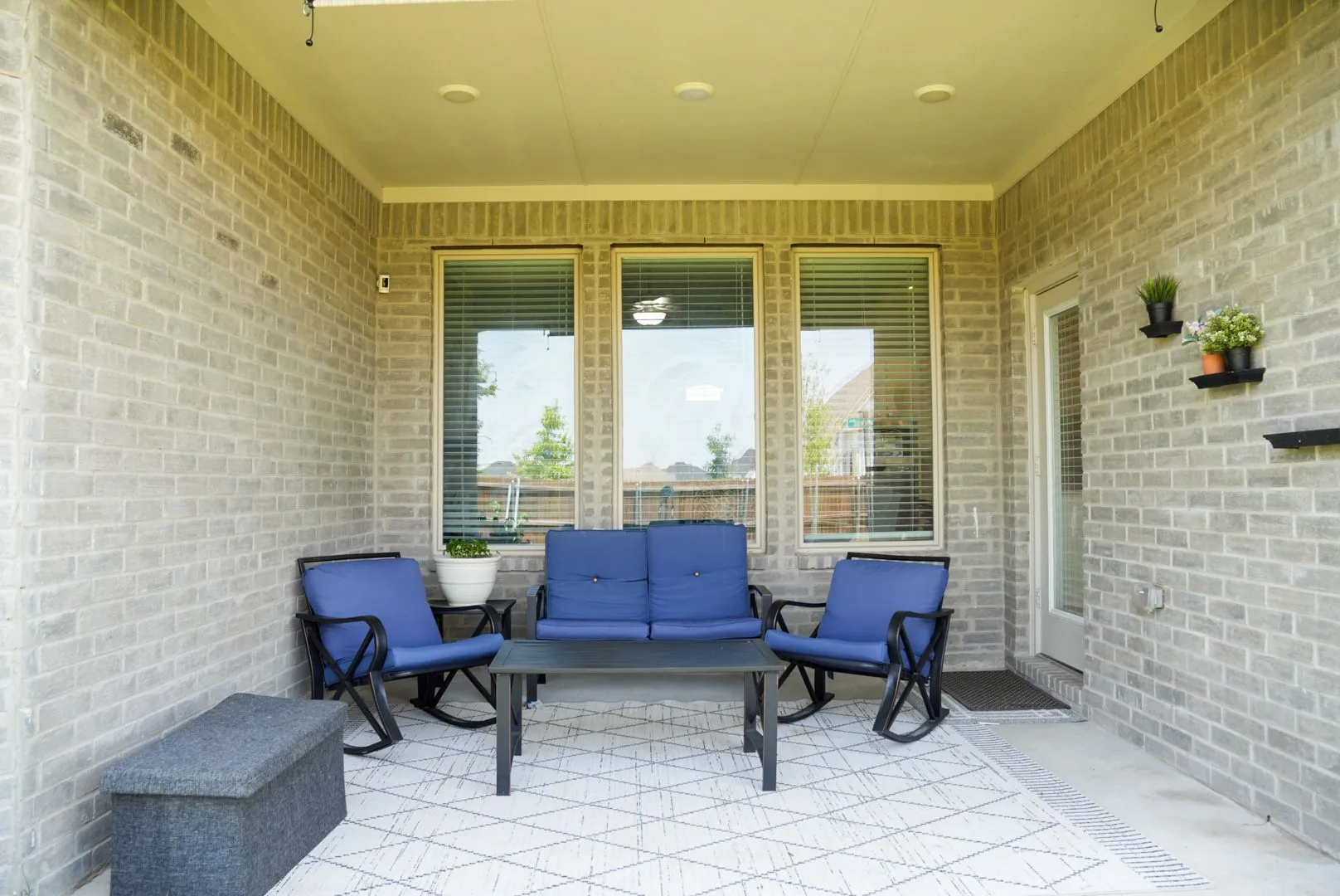 Covered patio with light-colored brick exterior, three large windows, and a glass-paneled door