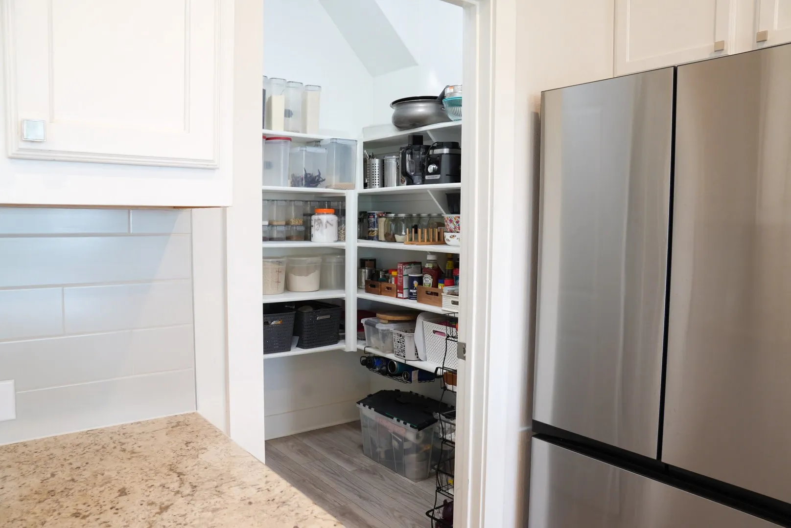 White shaker cabinetry, light-toned countertop, subway tile backsplash, stainless steel refrigerator, and a walk-in pantry with wood-finish flooring