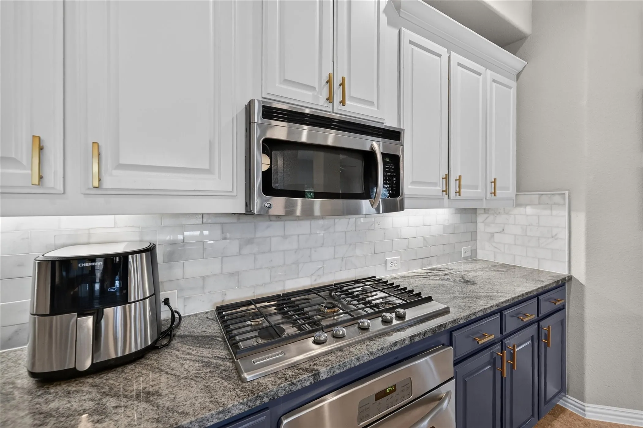 Two-tone cabinetry with gold-toned hardware, stainless steel built-in microwave, gas cooktop, stone countertops, and white subway tile backsplash