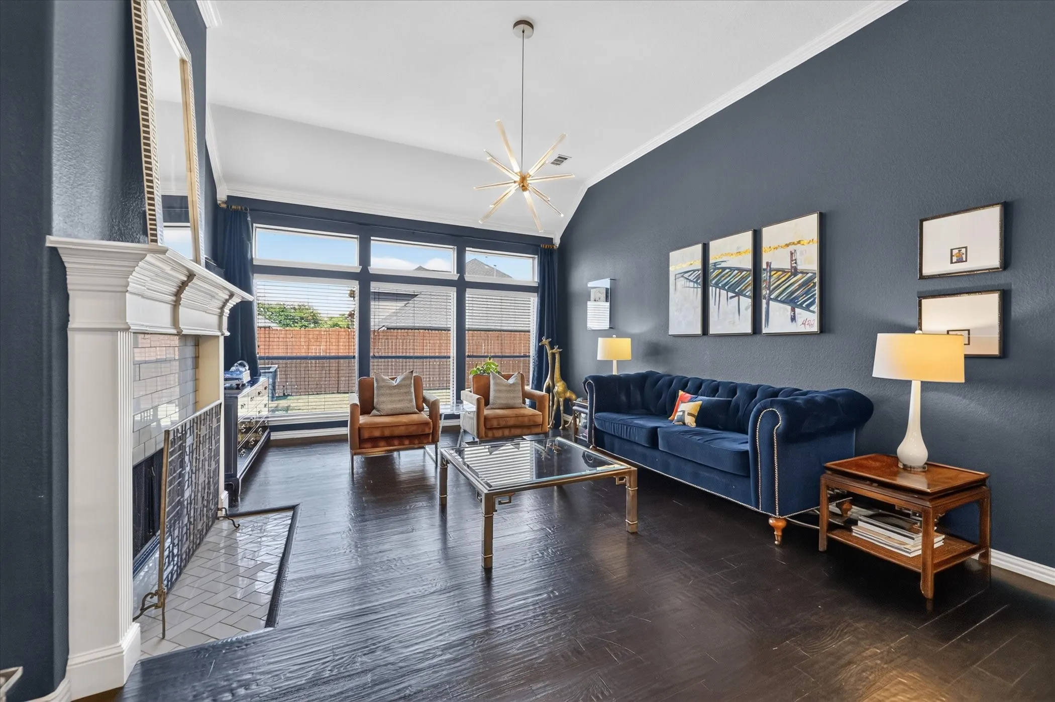 Vaulted ceiling with an angular chandelier, dark wood-finish flooring, a white mantel fireplace with a tiled hearth, and a wall of windows