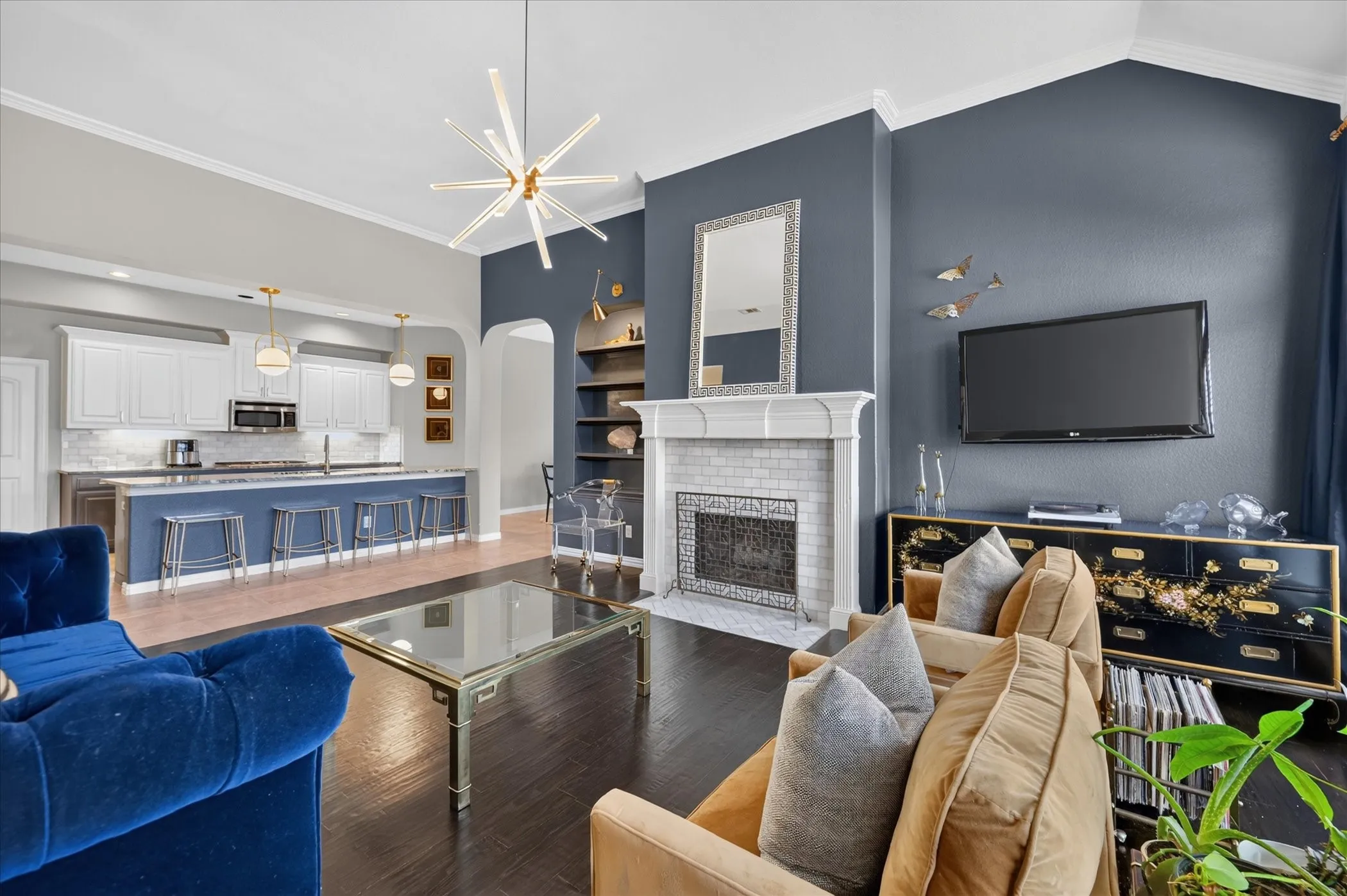 Vaulted ceilings with crown molding, a fireplace with a tile surround and decorative mantel, built-in shelving, an open-concept kitchen with pendant lighting, and wood-finish flooring