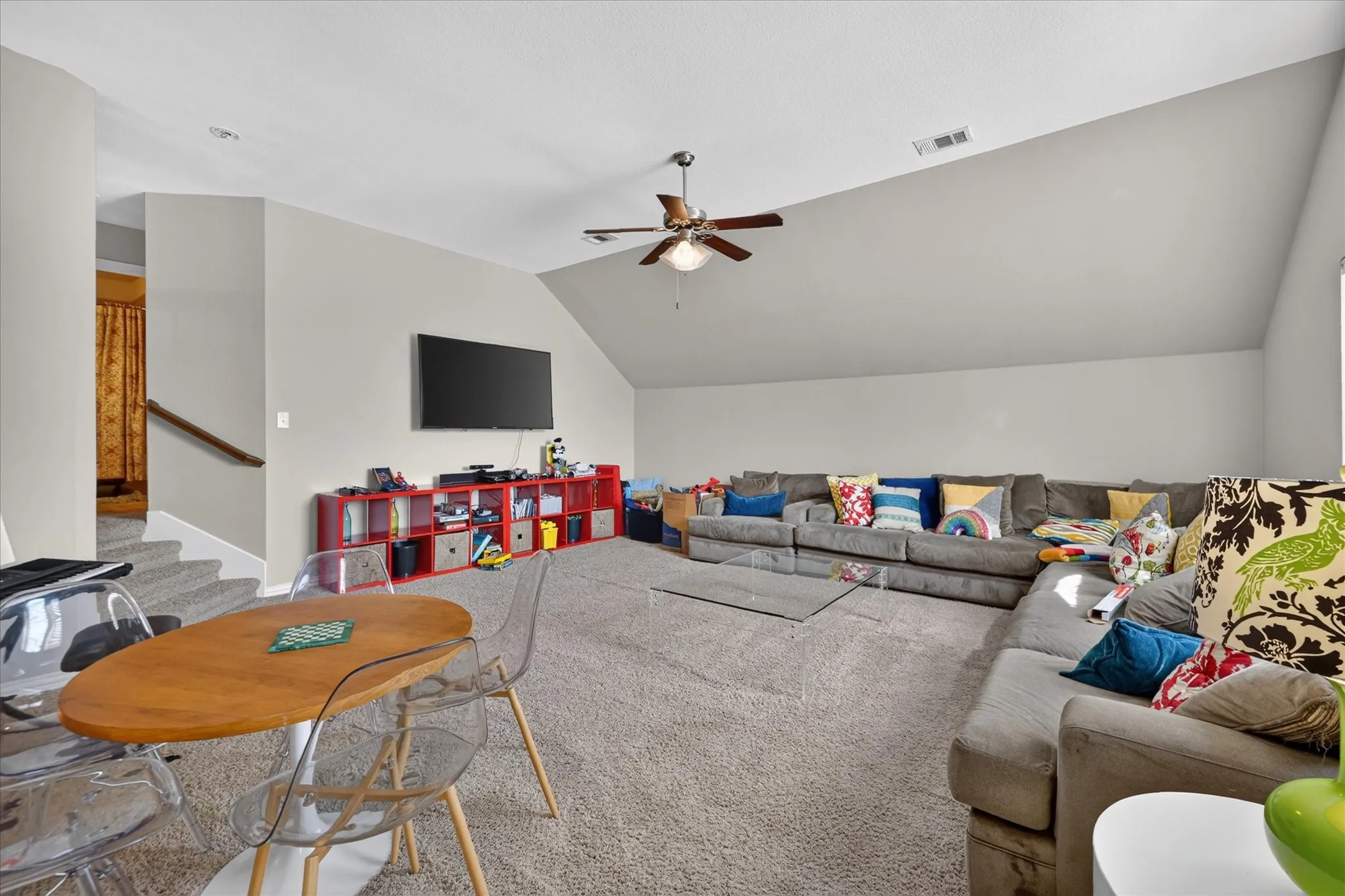 Carpeted room featuring a sloped ceiling, ceiling fan, wall-mounted television, and a visible staircase with a wooden handrail