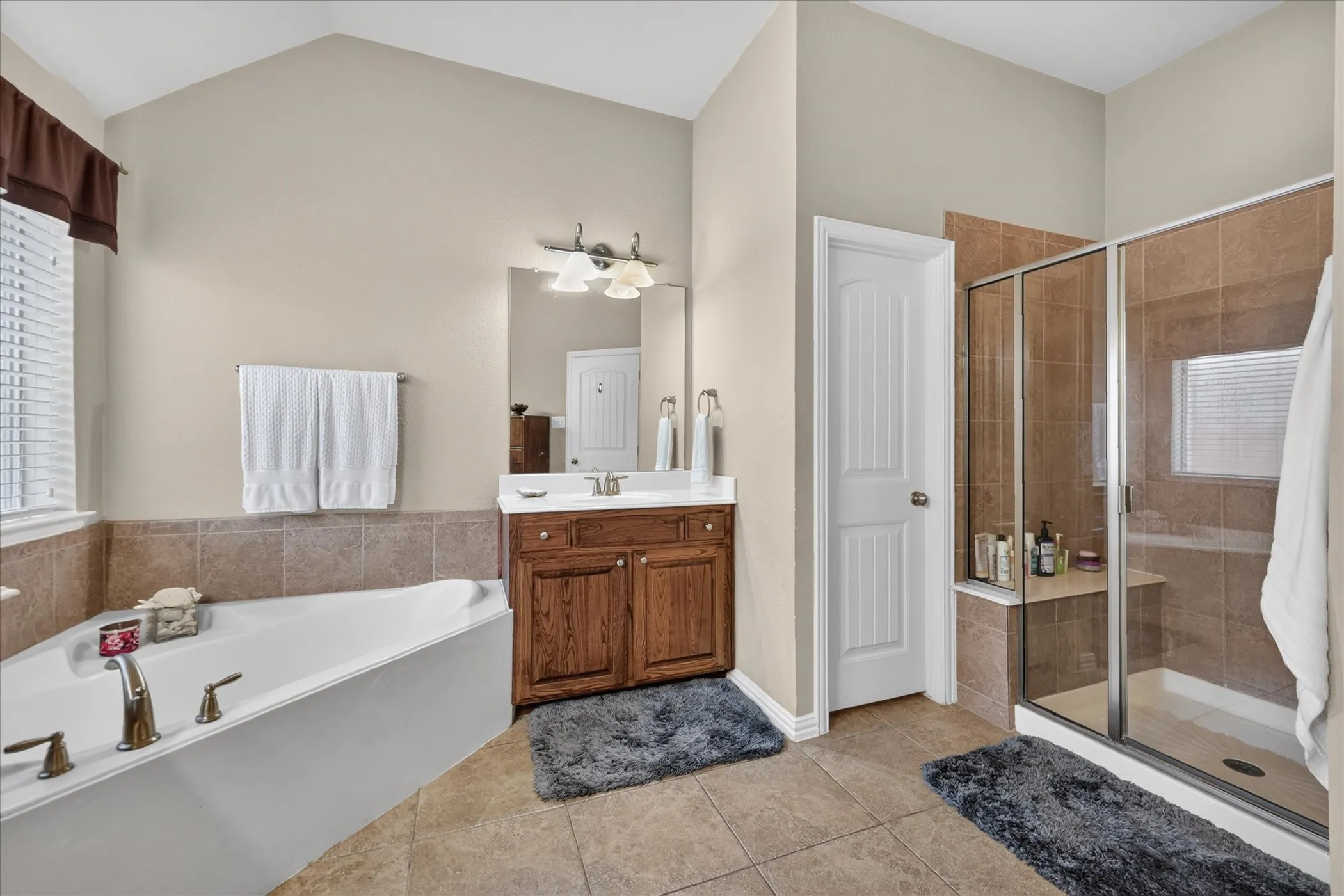 Vaulted ceiling, corner soaking tub with deck-mounted fixtures, wood-finish vanity with countertop sink, glass-enclosed shower with tiled walls, and ceramic tile flooring