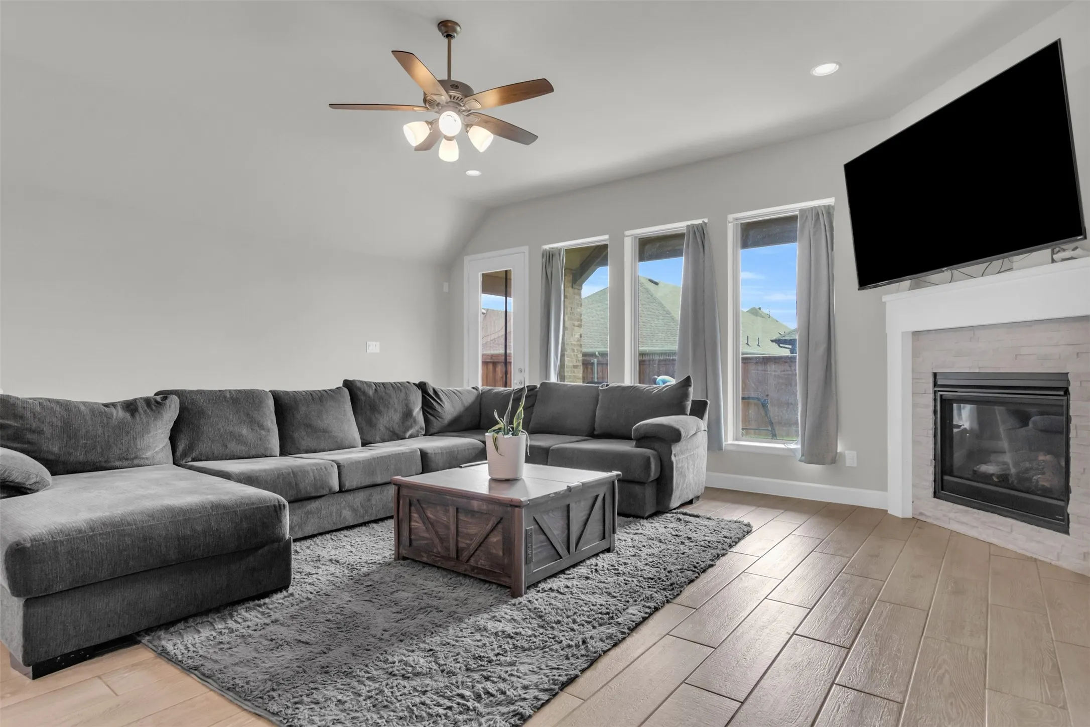 Living area with light wood-style flooring, a glass covered fireplace, a ceiling fan, vaulted ceiling, and recessed lighting