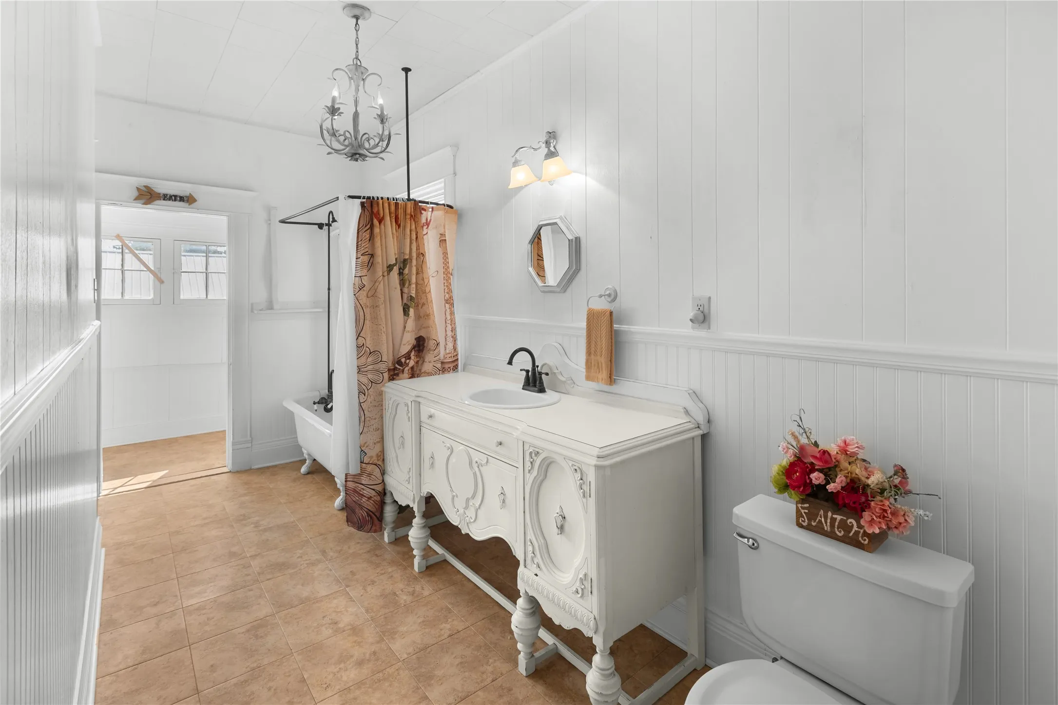 White paneled walls with wainscoting, decorative vanity with an integrated sink, clawfoot tub with a shower surround, tiled flooring, and a chandelier fixture