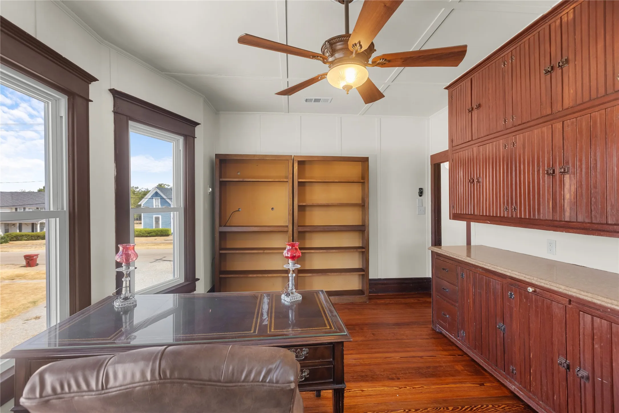 Wood-finish flooring, built-in cabinetry with a countertop, two windows with dark trim, and a ceiling fan with integrated lighting