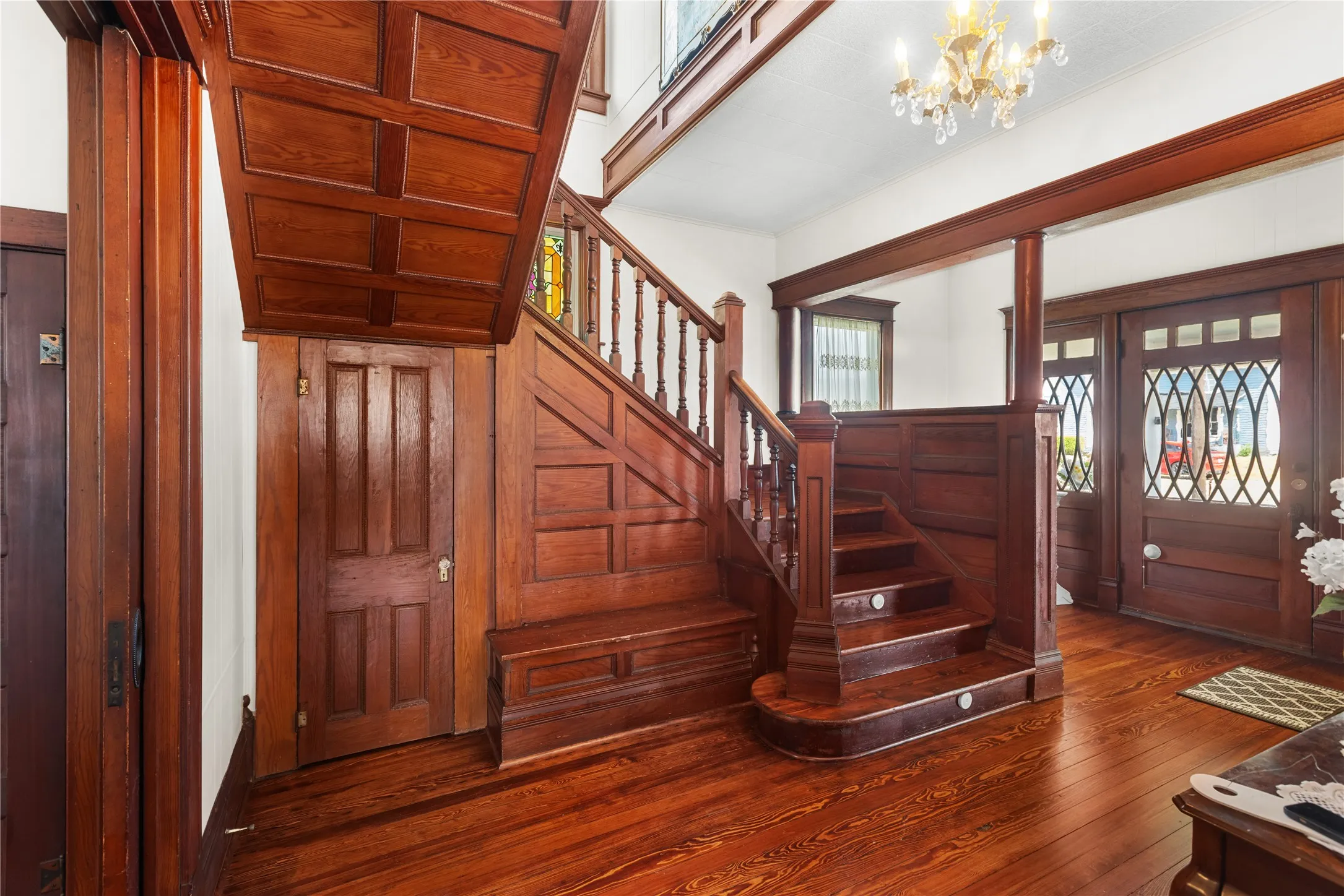 Hardwood flooring with a detailed staircase, paneled walls, decorative entry doors, and an overhead chandelier