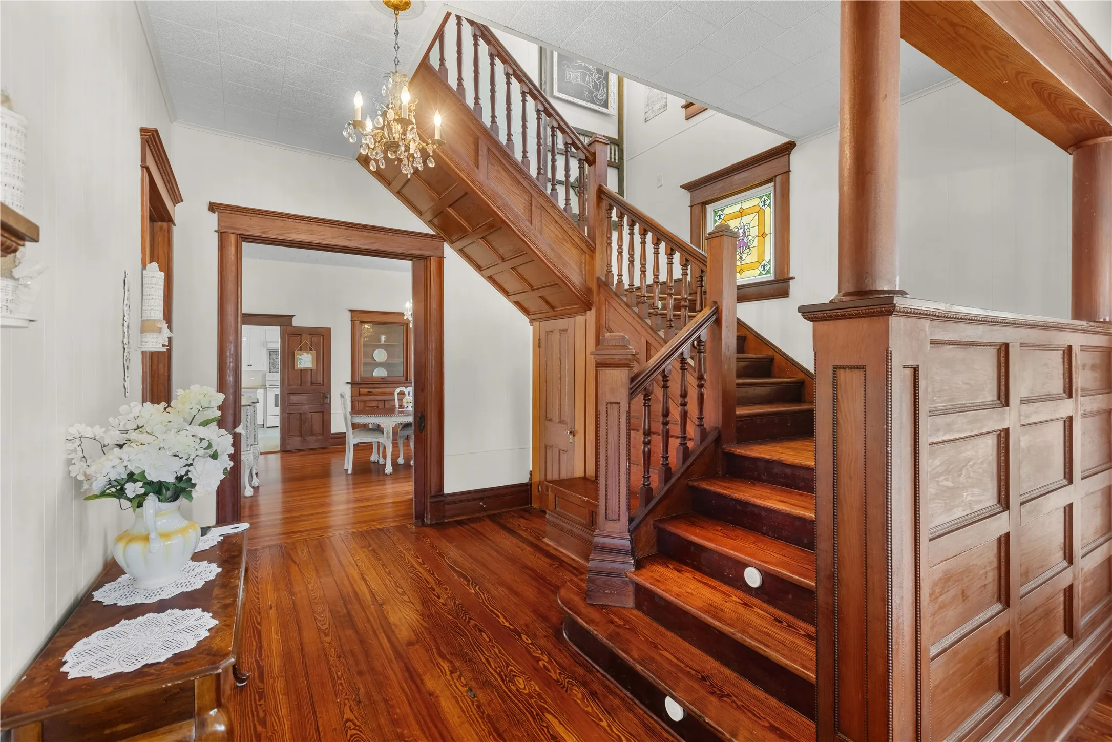Hardwood flooring throughout, a grand wooden staircase with decorative balusters, a crystal chandelier, intricate wood paneling, and an arched doorway framing another room