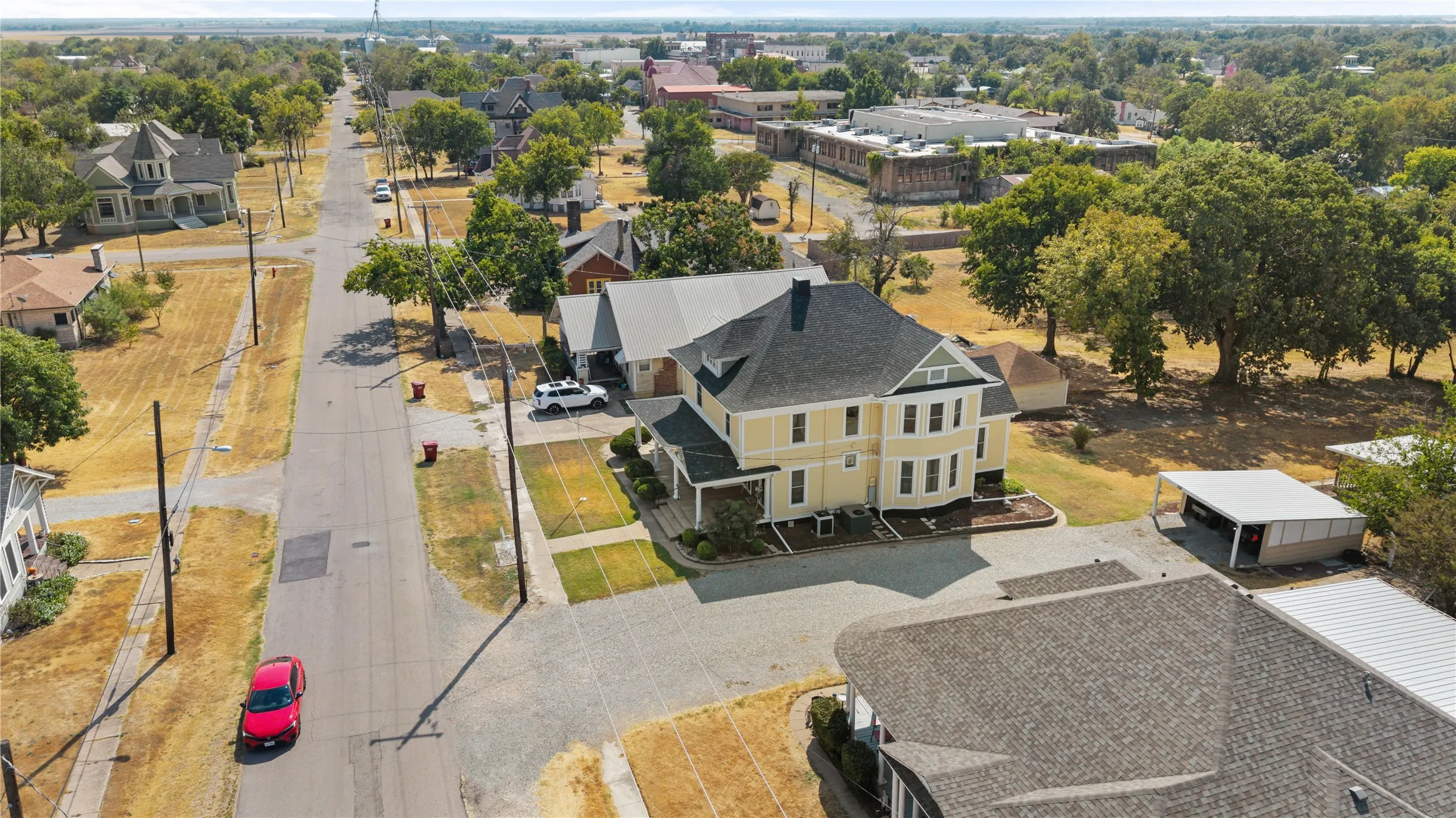 Yellow exterior with white trim, dark shingle roof, established landscaping, and a paved driveway