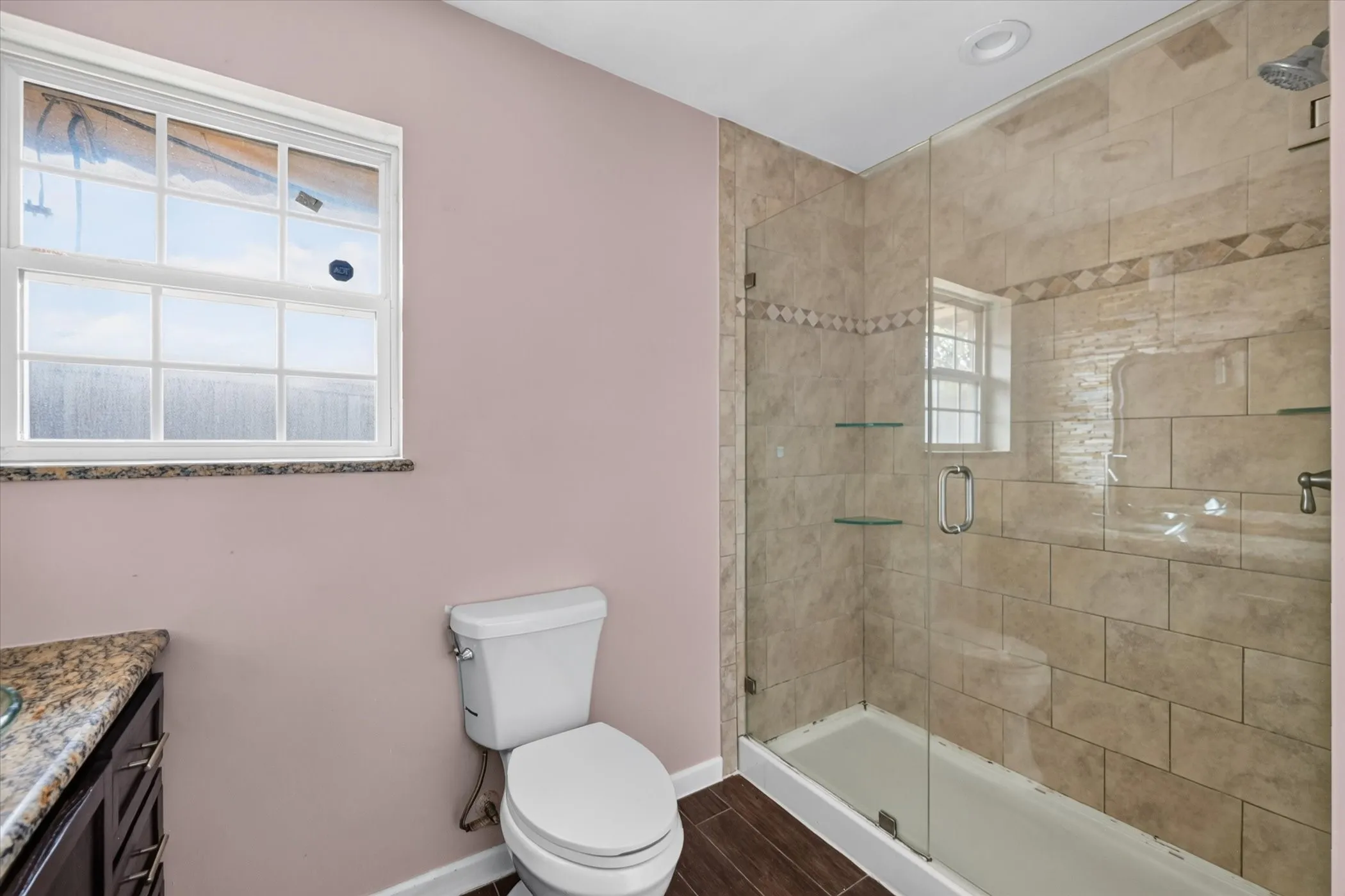 Glass-enclosed shower with tile surround and integrated shelving, dark wood-finish flooring, and a window with a stone sill