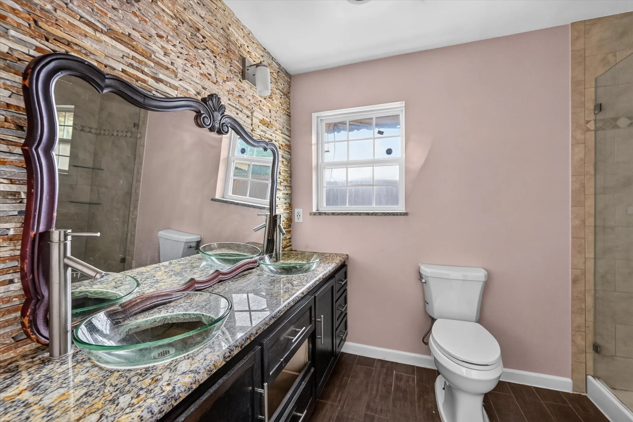 Stacked stone accent wall with a large decorative mirror, double vessel sinks, granite countertop vanity, and wood-finish flooring