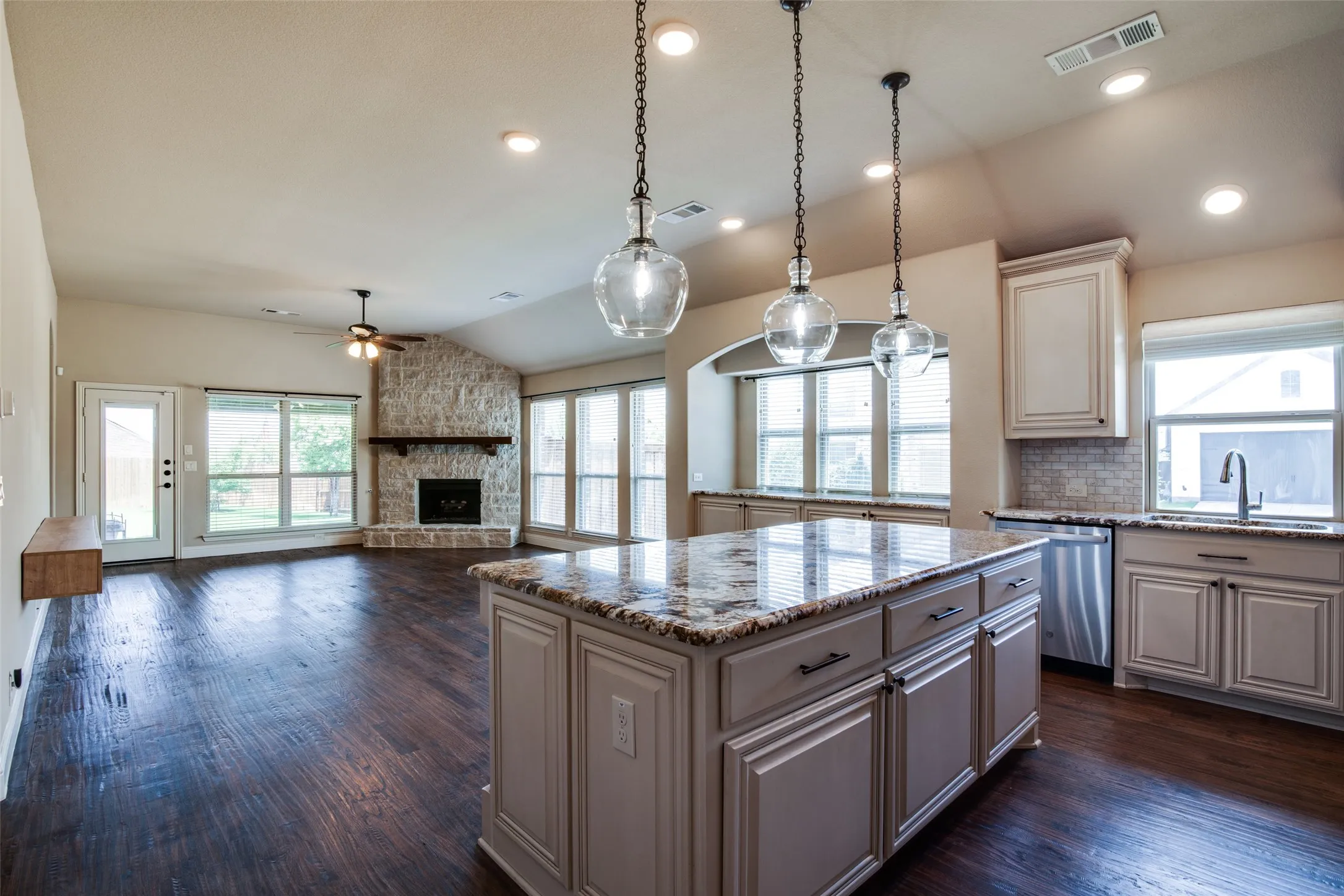 Wood-finish flooring throughout, a stone fireplace, recessed lighting, and a kitchen island with pendant lighting