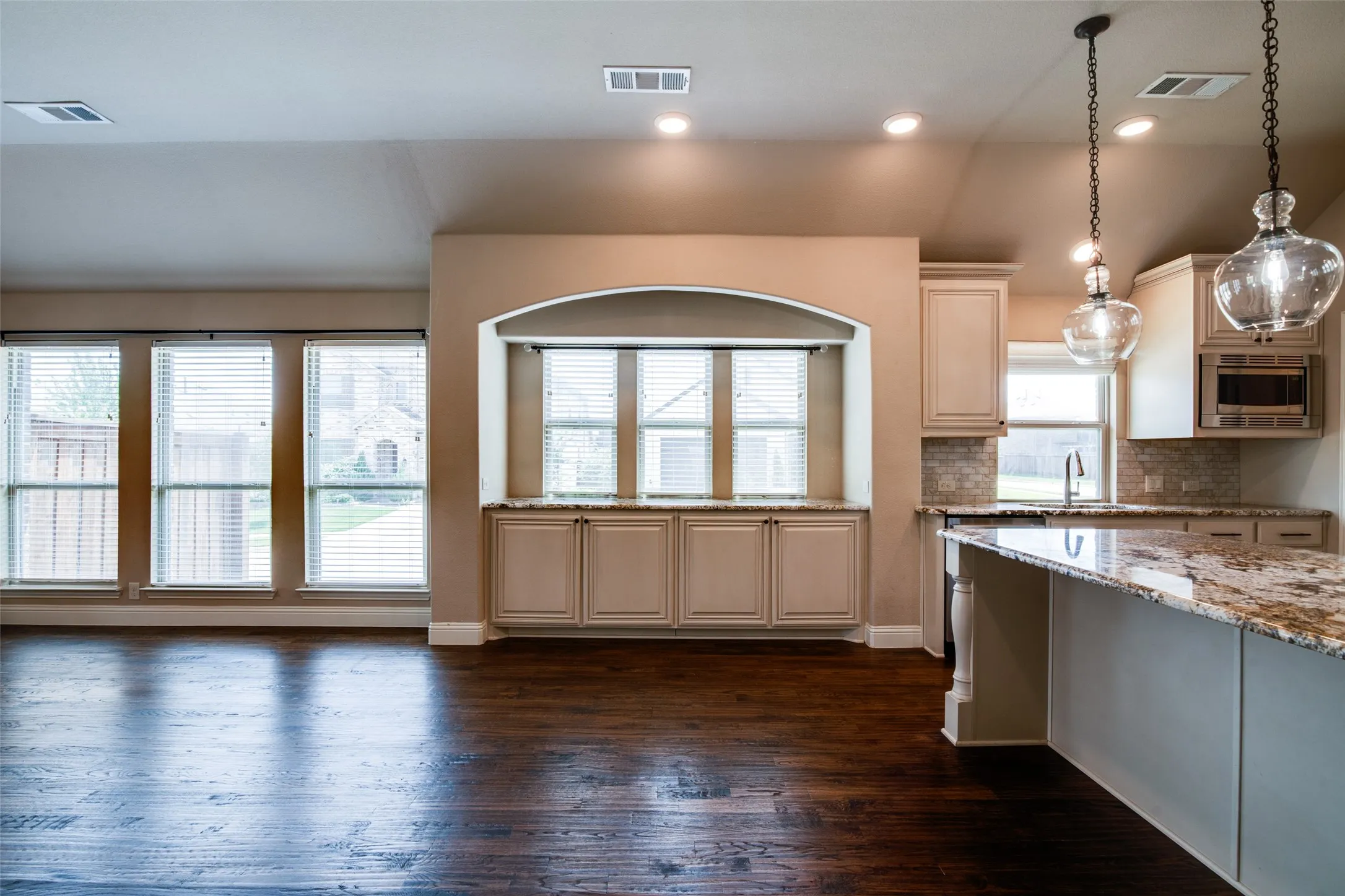 Open-concept living and kitchen featuring dark wood-finish flooring, custom cabinetry with arched architectural detailing, granite countertops, stainless steel appliances, and pendant lighting
