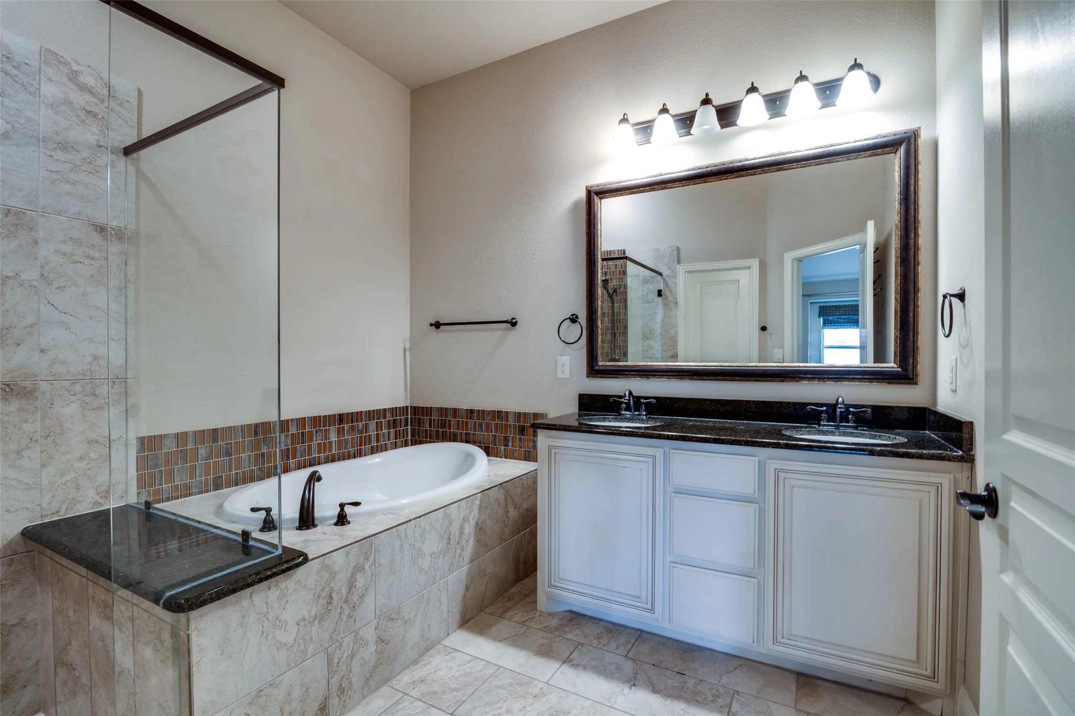 Dual sink vanity with dark countertop, large framed mirror, built-in soaking tub, glass-enclosed shower, and tiled flooring