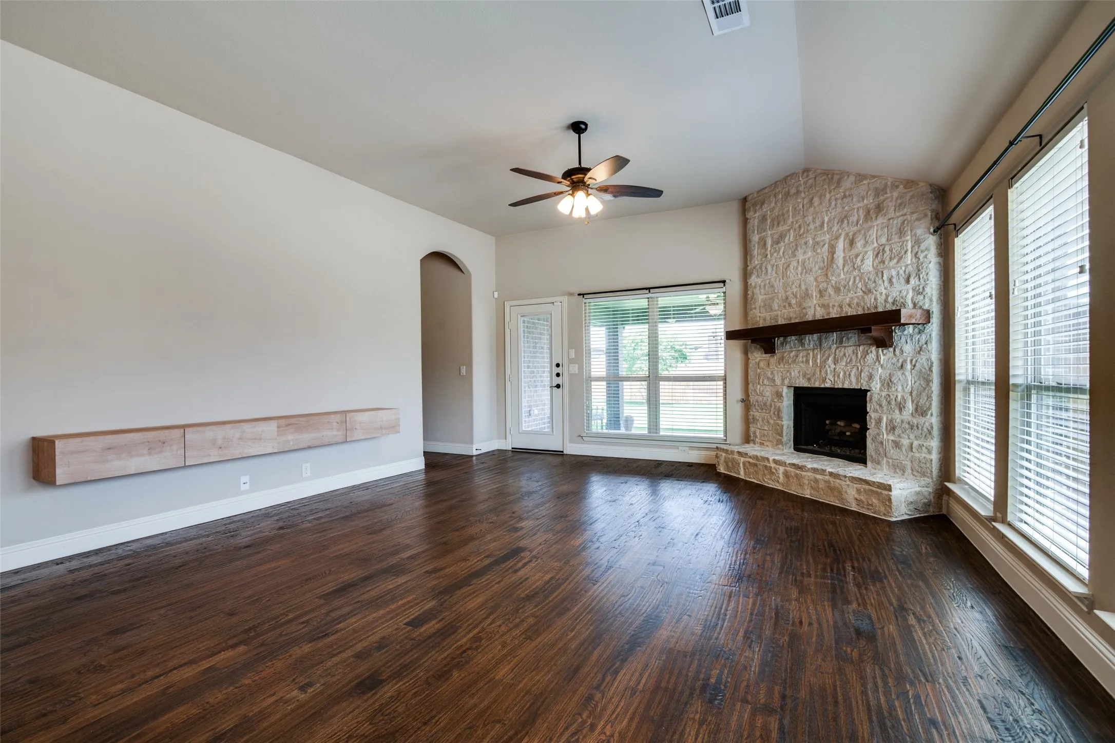 Spacious living area showcasing rich wood-finish flooring, ceiling fan with integrated lighting, and stone fireplace with wooden mantel