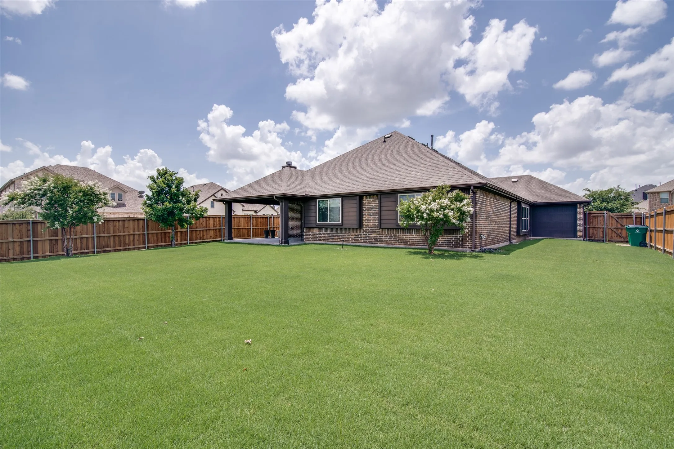 Expansive backyard featuring a manicured lawn, wood fencing, covered patio, and brick exterior