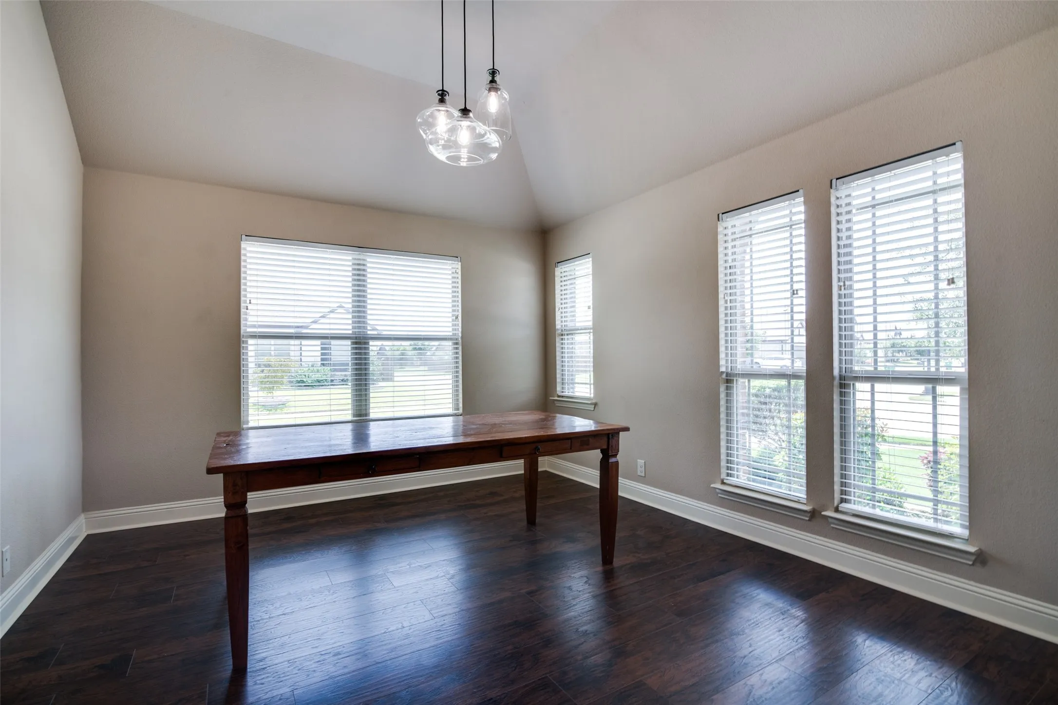 Vaulted ceiling, wood-finish flooring, multiple windows with blinds, and a contemporary pendant light fixture
