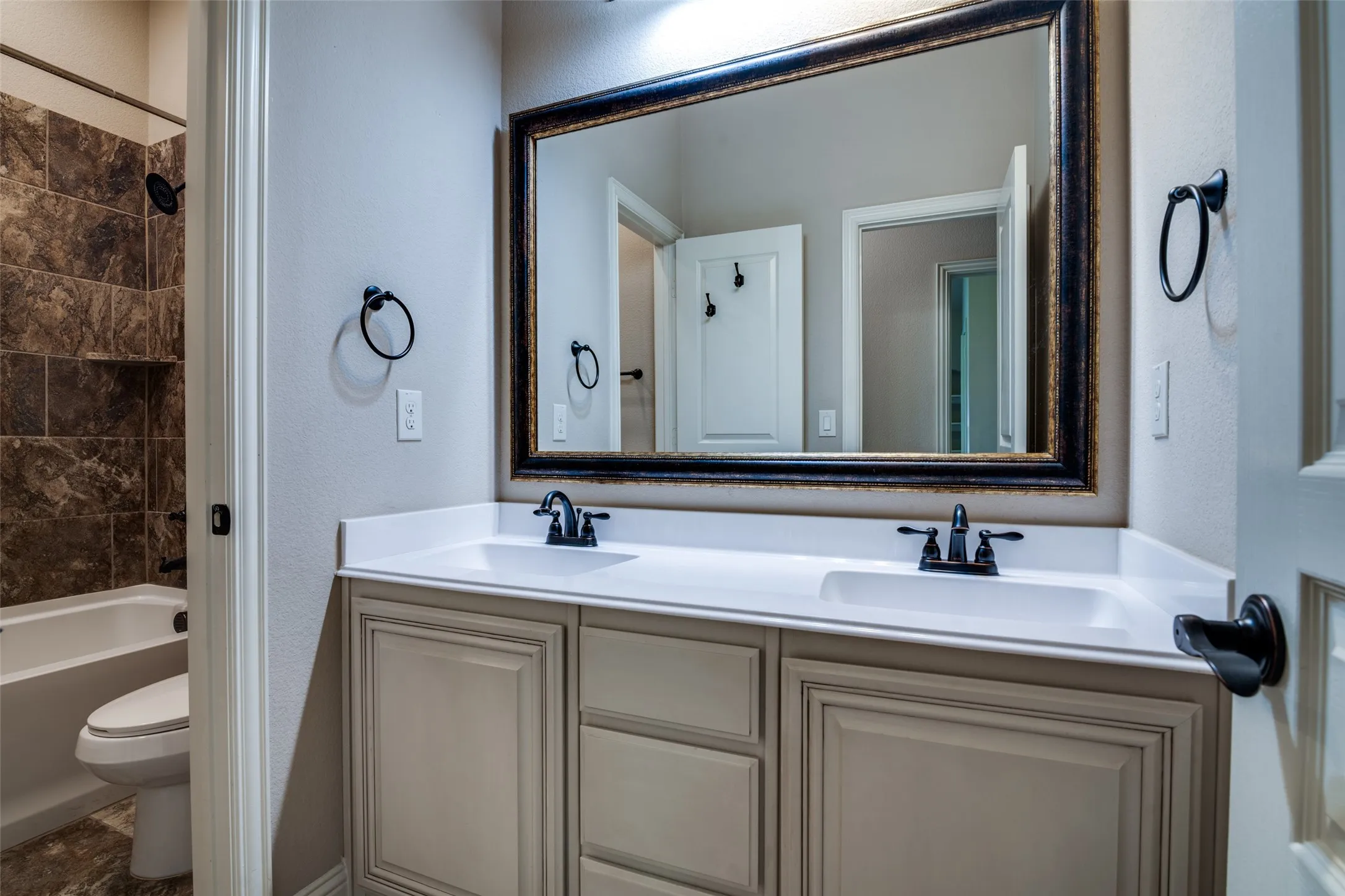 Dual vanity with white countertop and integrated sinks, framed mirror, matte black fixtures, built-in bathtub with shower, and tiled surround