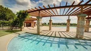 Resort-style outdoor entertaining space featuring a sparkling saltwater pool with sun-lounging deck, wood-lattice pergola overhead providing dappled shade, and stone column supports
