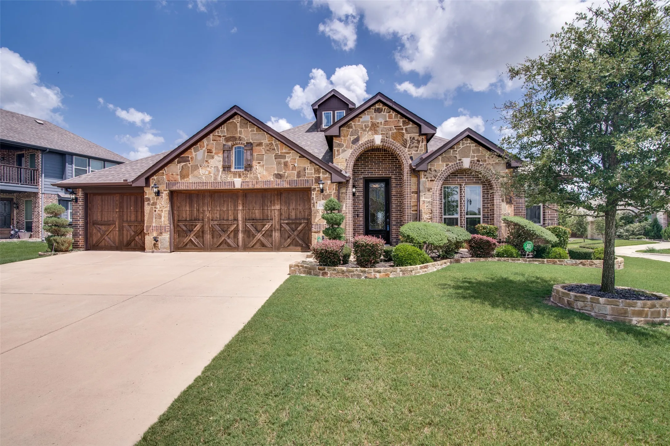 Stone and brick exterior with arched entryway, wood-finish garage doors, and established landscaping