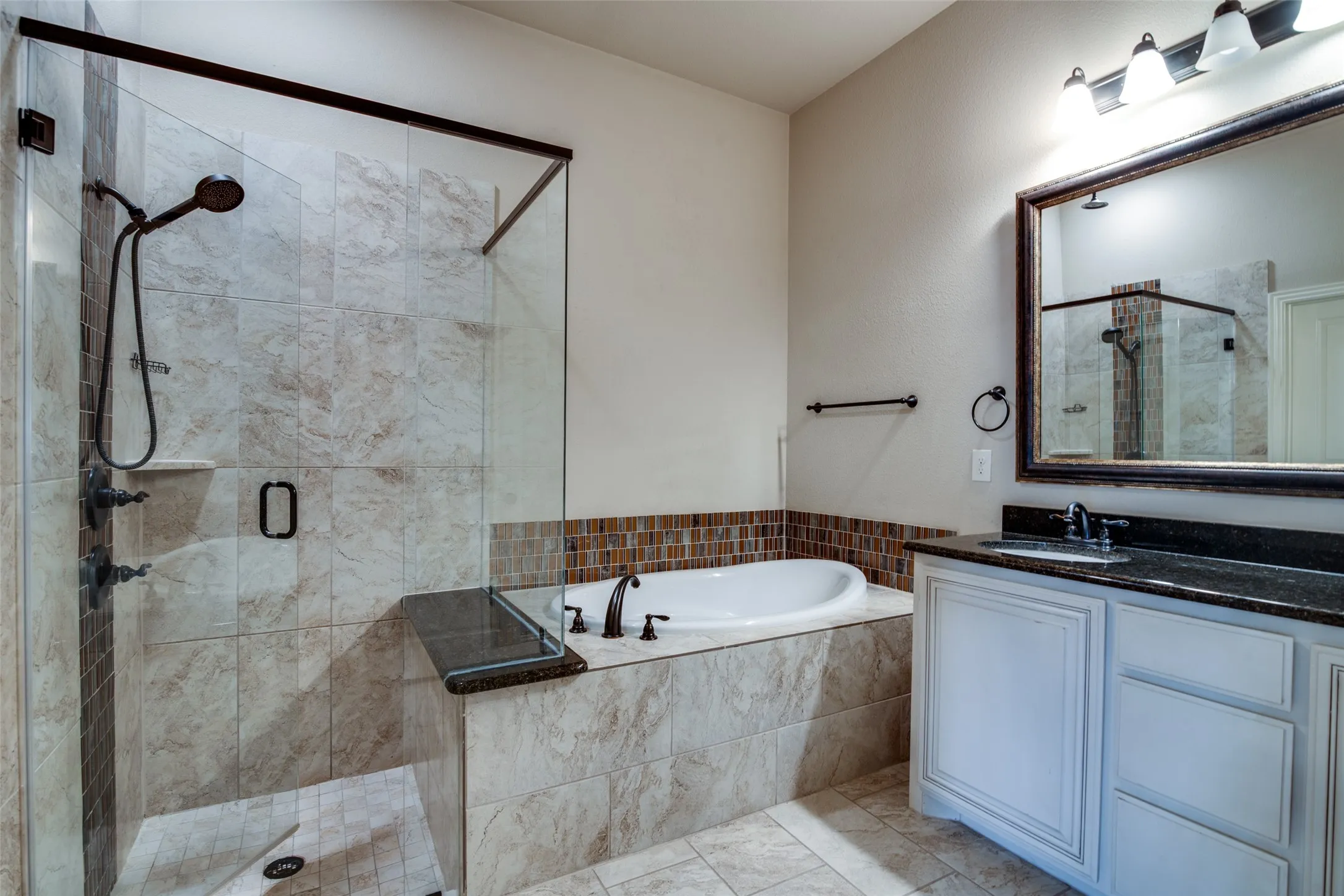 Glass-enclosed shower with tile surround and integrated shelving, soaking tub with tile surround and mosaic accent, single vanity with dark countertop and white cabinetry, wall-mounted mirror with decorative frame, and tile flooring