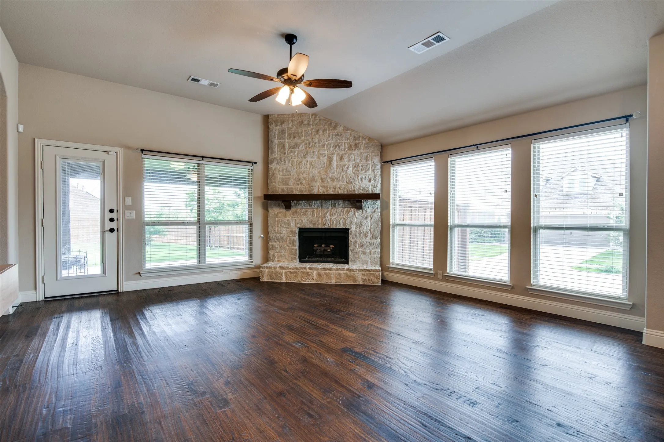 Vaulted ceiling with a ceiling fan, stone-clad corner fireplace with a wood mantle, rich wood-finish flooring, and multiple windows