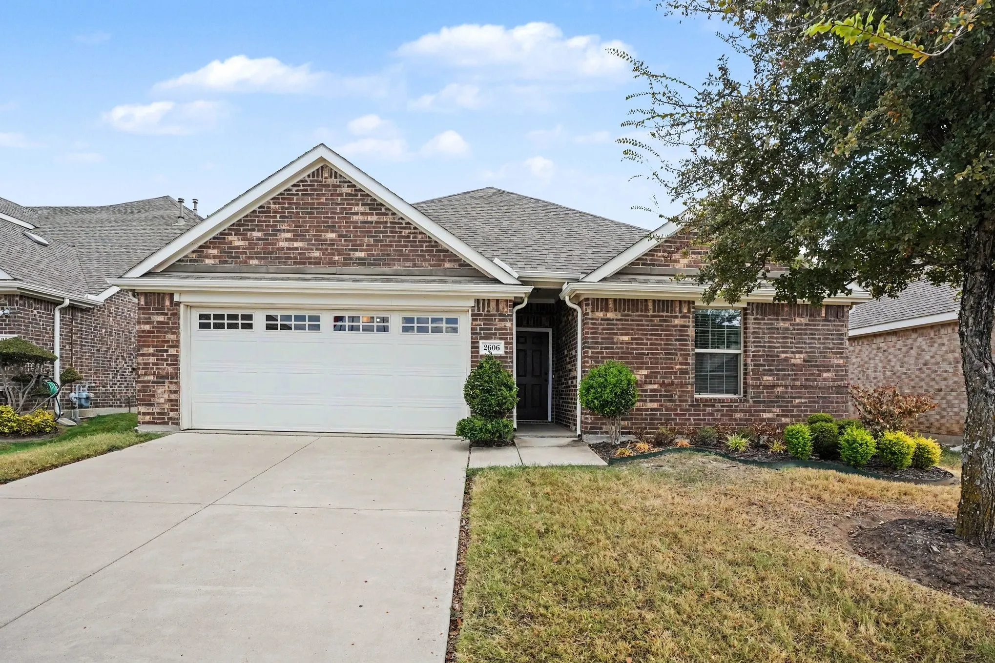 Brick exterior with a gabled roofline, attached garage with decorative windows, concrete driveway, and established front landscaping