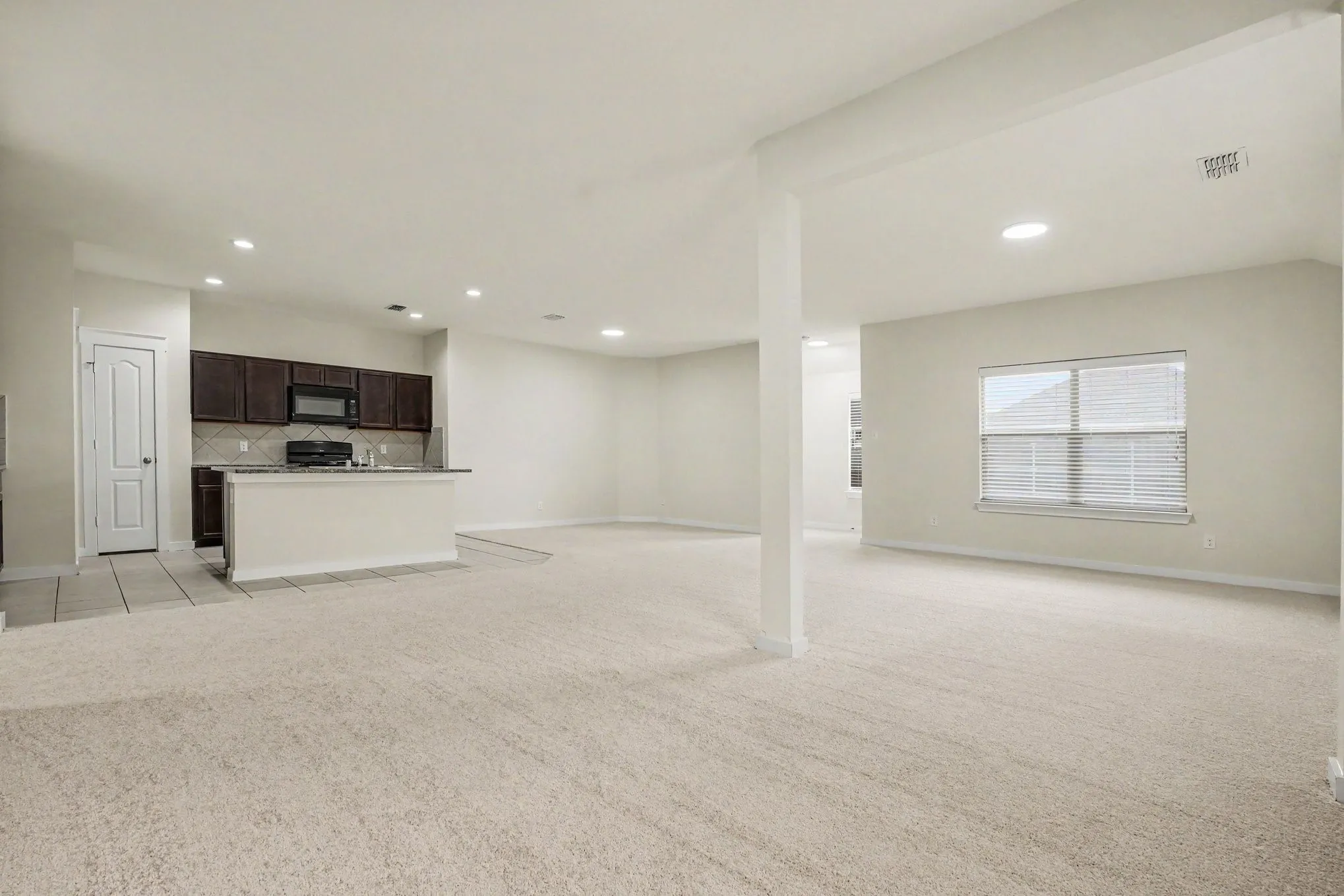Open-concept living area with neutral carpeting, recessed lighting, and a kitchen featuring dark wood cabinetry, a built-in microwave, and a breakfast bar