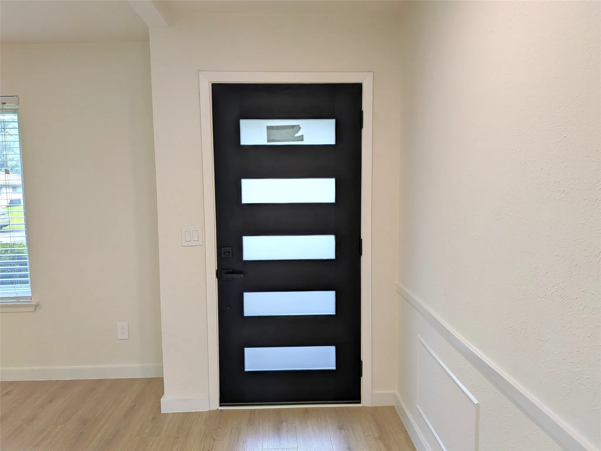 Contemporary entry door with frosted glass panels, matte black hardware, light wood-finish flooring, and decorative wainscoting