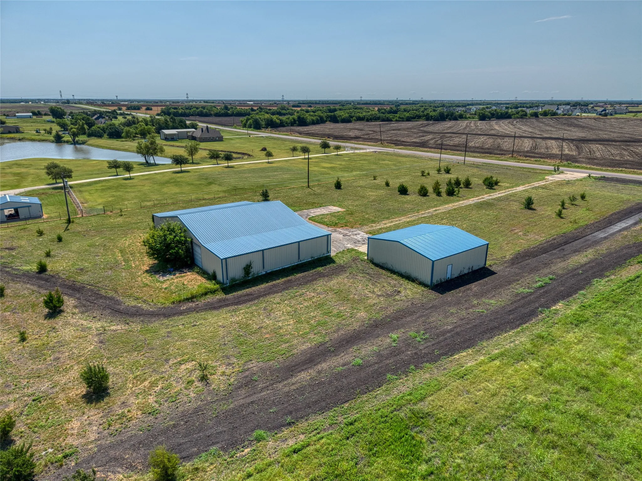 Expansive rural property featuring two large metal-frame agricultural structures with blue metal roofing, situated on manicured grounds with newly planted landscaping