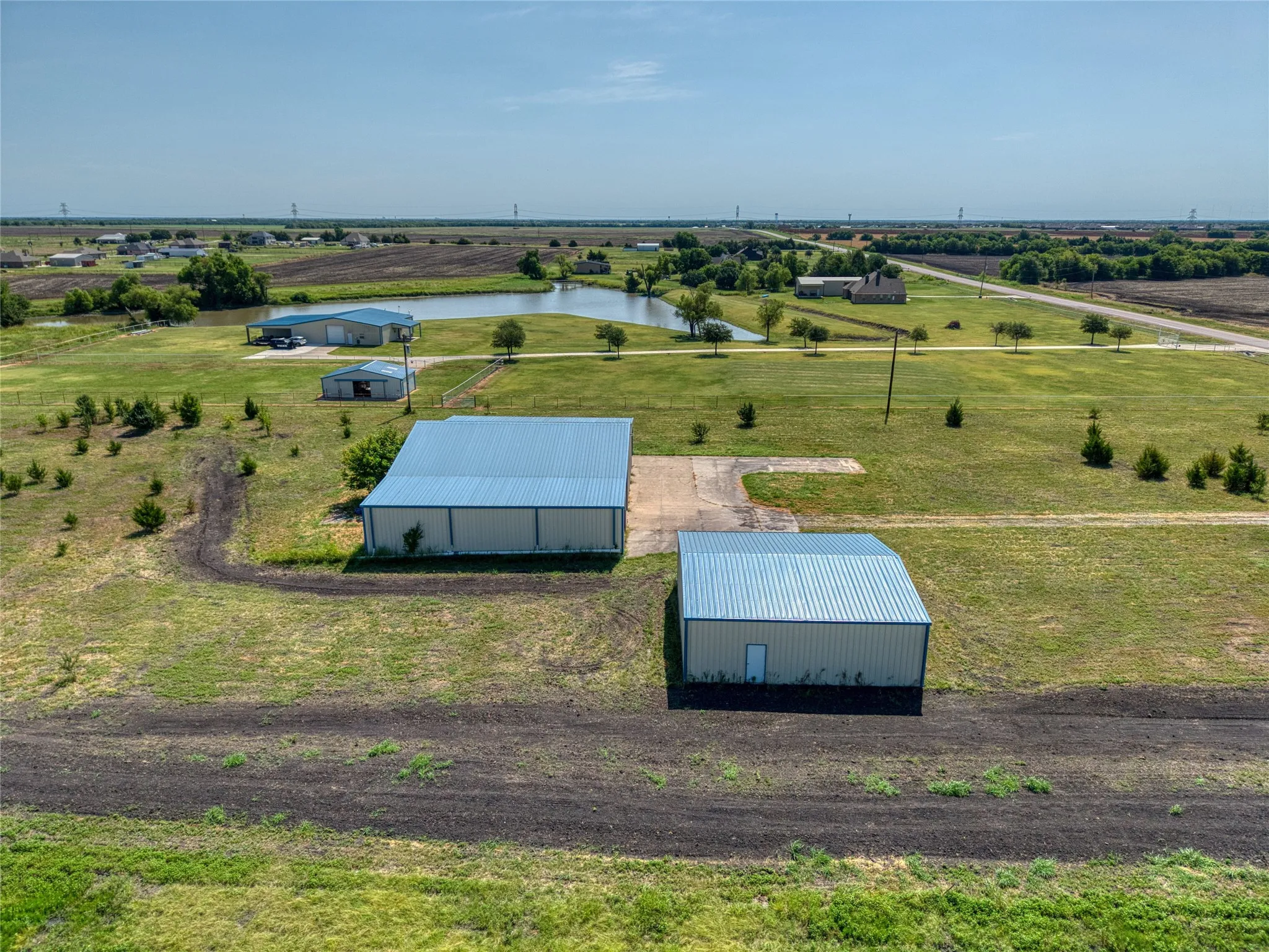 Two metal buildings with light blue roofs, one larger and one smaller, are set on a property featuring a pond, a concrete pad, and an expanse of green lawn