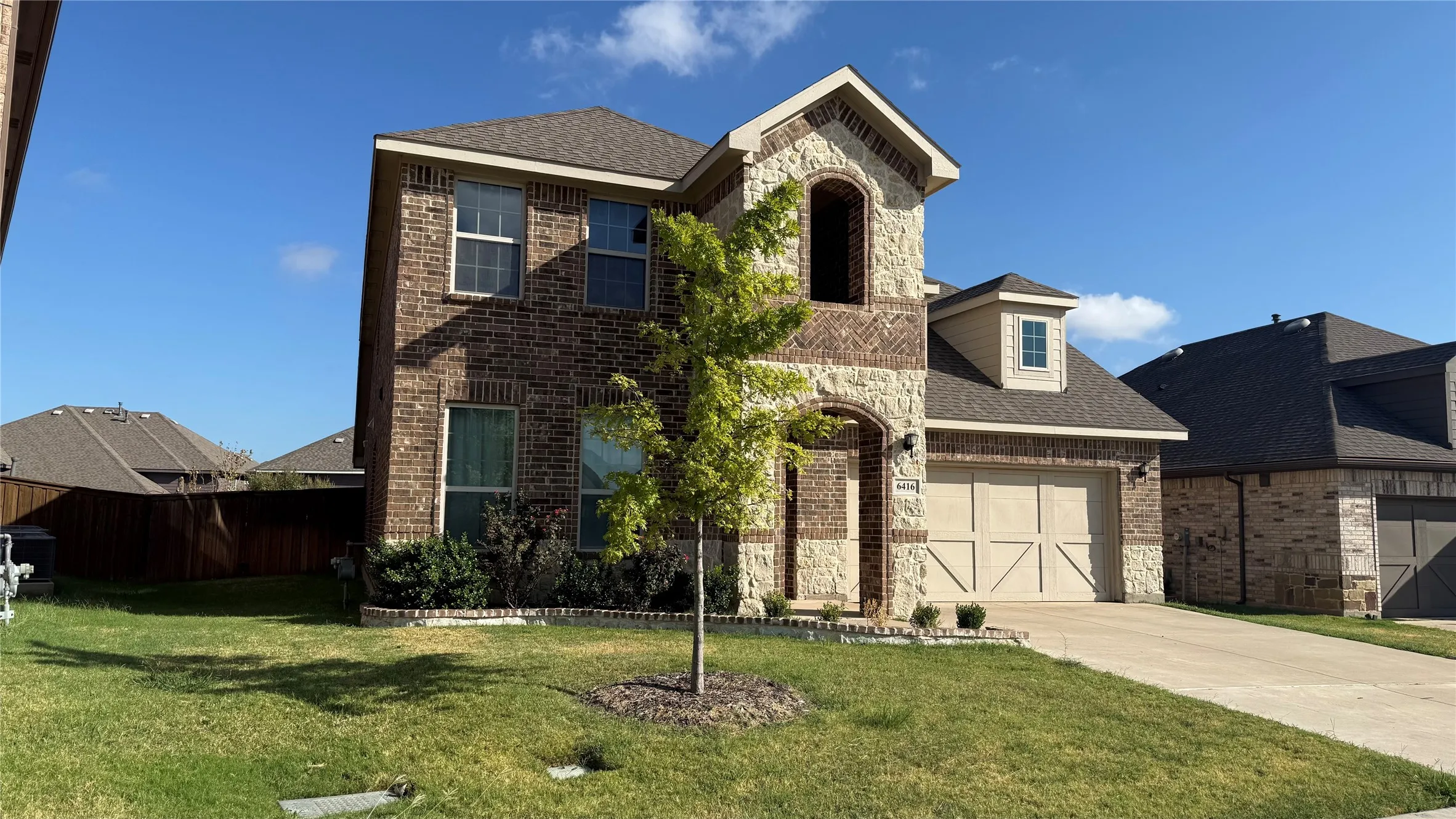 Brick and stone facade, dormer window, attached garage with decorative doors, paved driveway, and established front landscaping