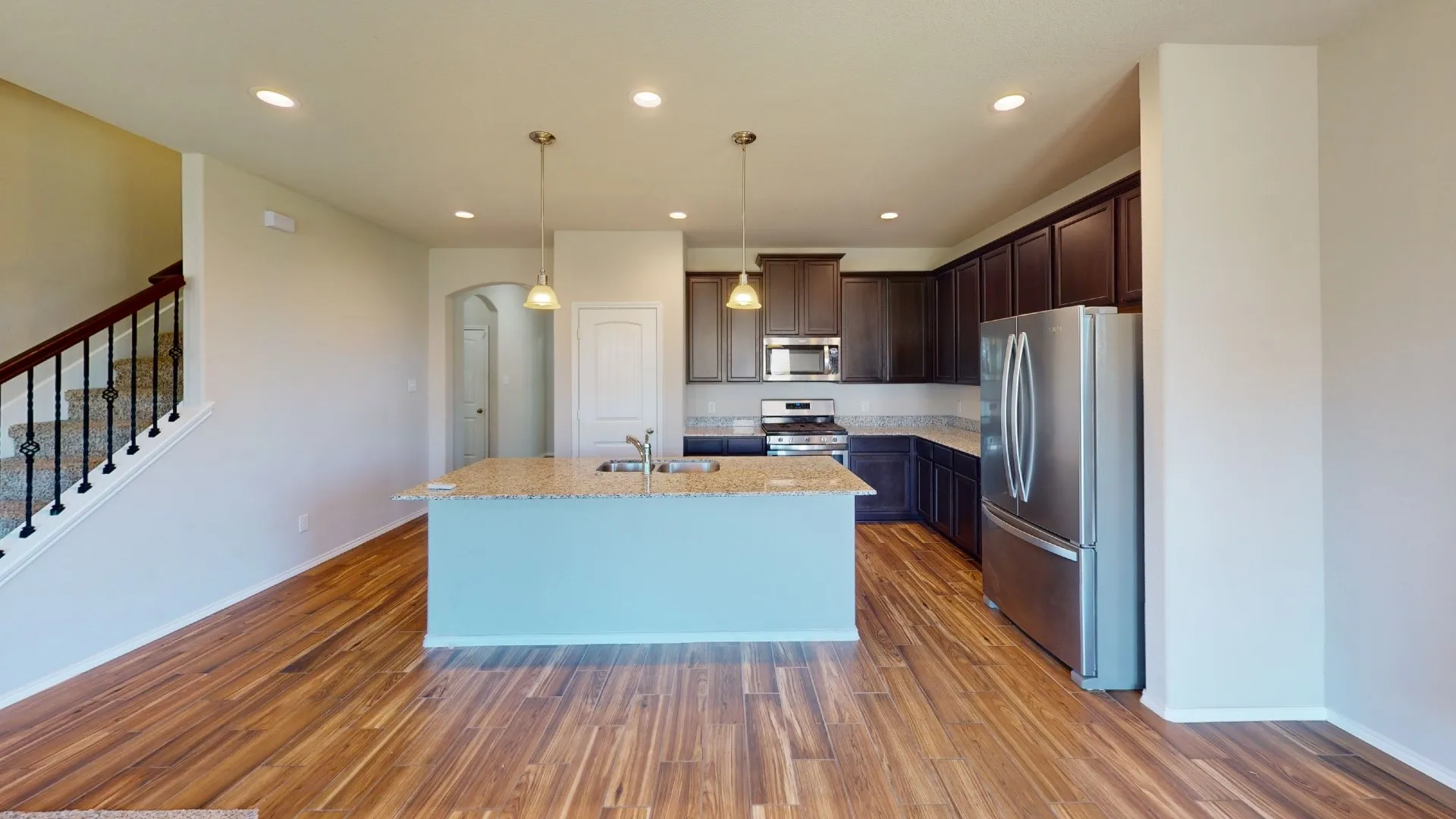 Open-concept kitchen and dining area featuring warm wood-finish flooring, a light blue island with granite countertop, stainless steel appliances, dark cabinetry, recessed ceiling lighting, and a bright, airy layout with abundant natural light